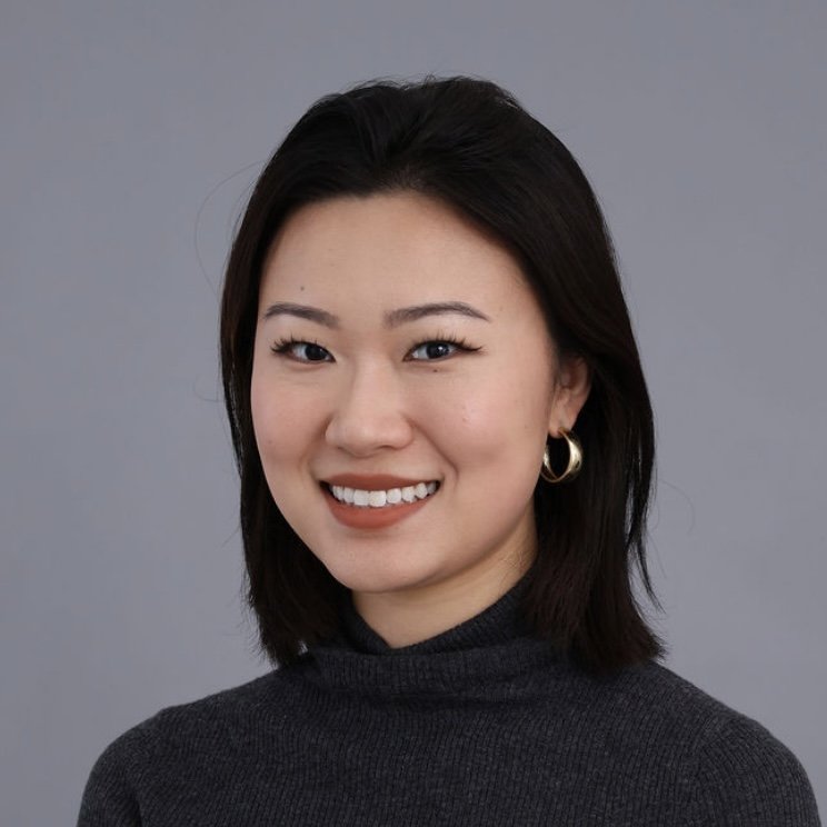 A young woman with dark hair styled in a shoulder-length bob, smiling and wearing a dark turtleneck sweater against a plain gray background.