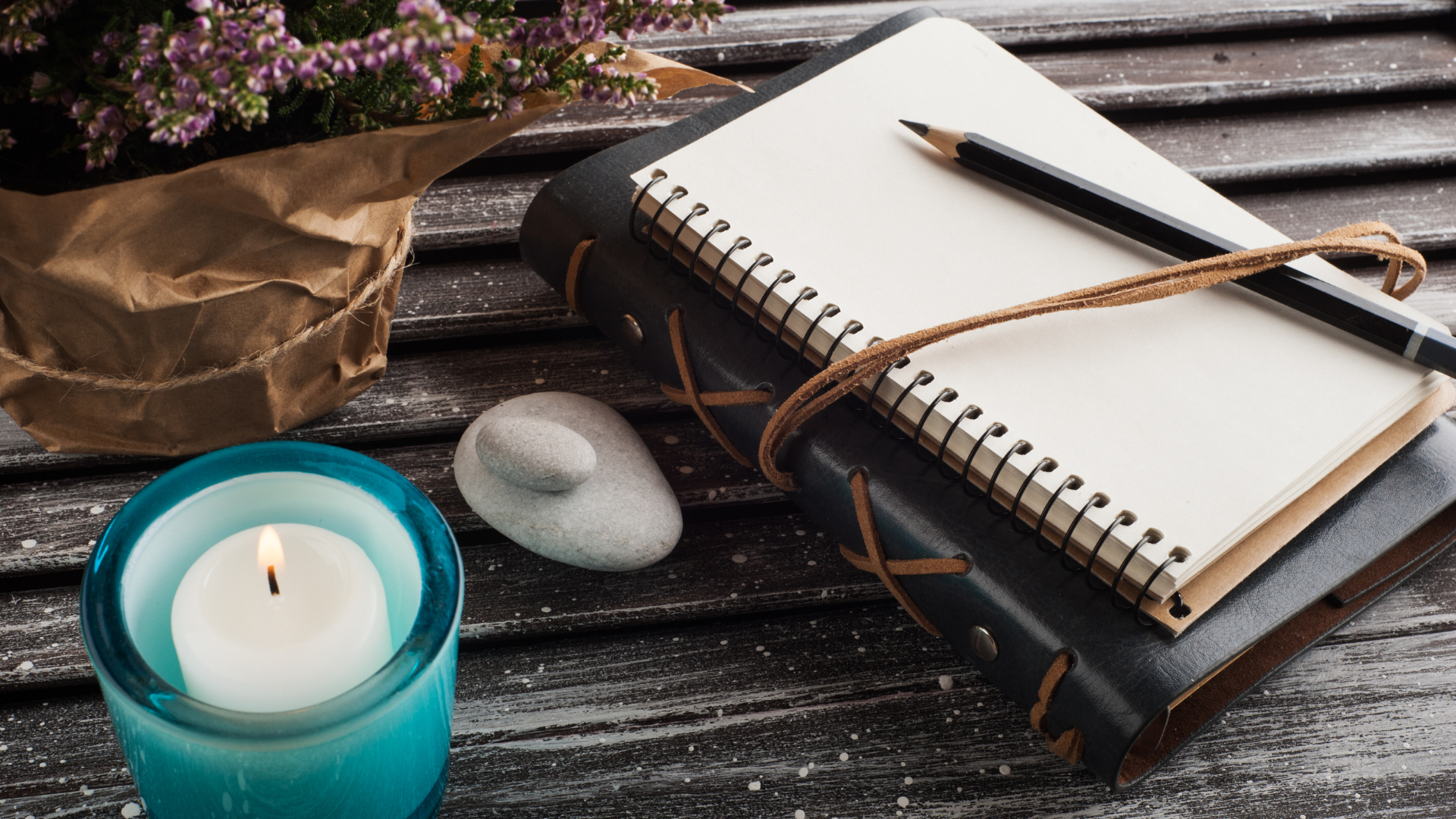 A wooden table with a lit candle in a blue holder, a stack of blank notebooks with a black pen, a gray stone, and a bouquet of purple flowers wrapped in brown paper.