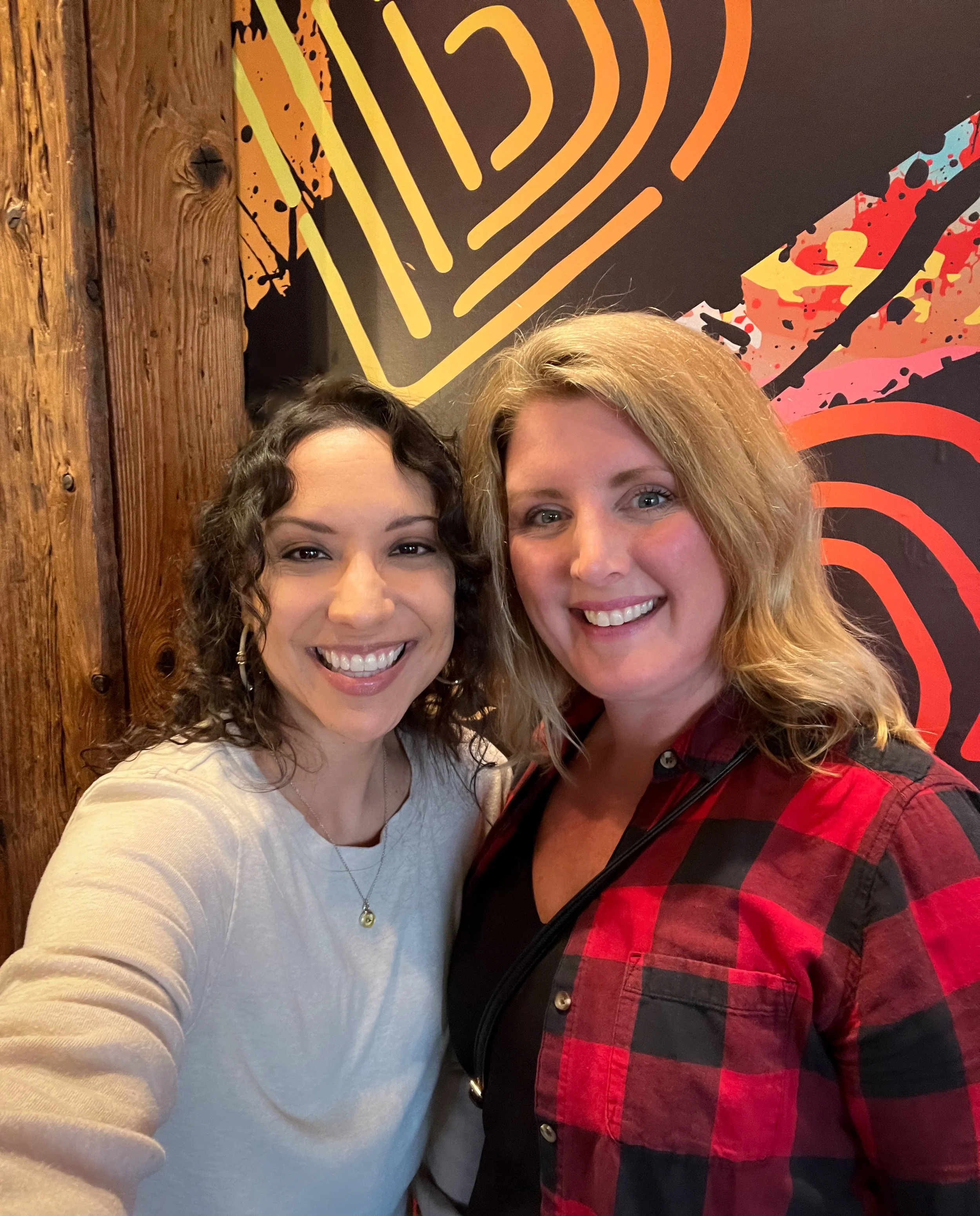 Two women taking a selfie in front of a colorful wall with abstract designs. The woman on the left has curly dark hair and is wearing a white top, while the woman on the right has wavy blonde hair and is wearing a red and black checkered shirt.