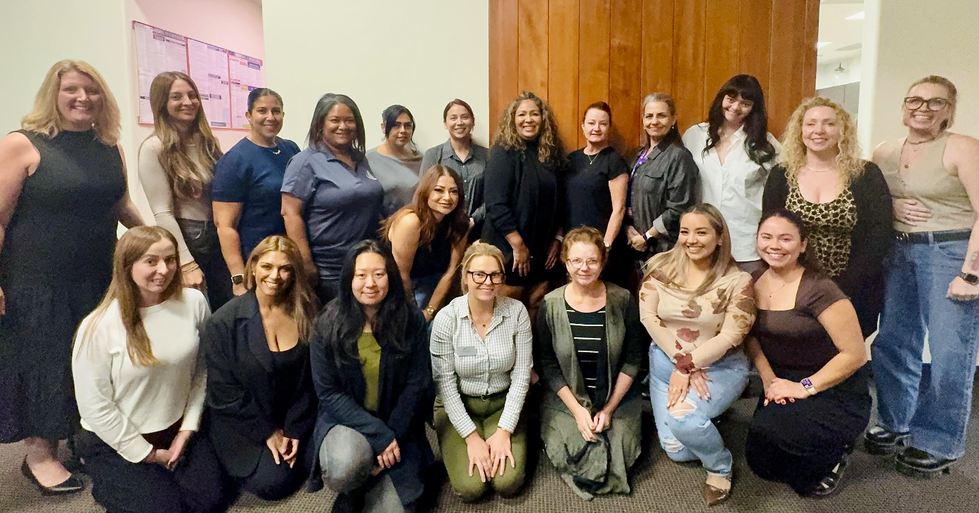 Group of women gathered indoors for a group photo, smiling and dressed in casual and business casual attire.