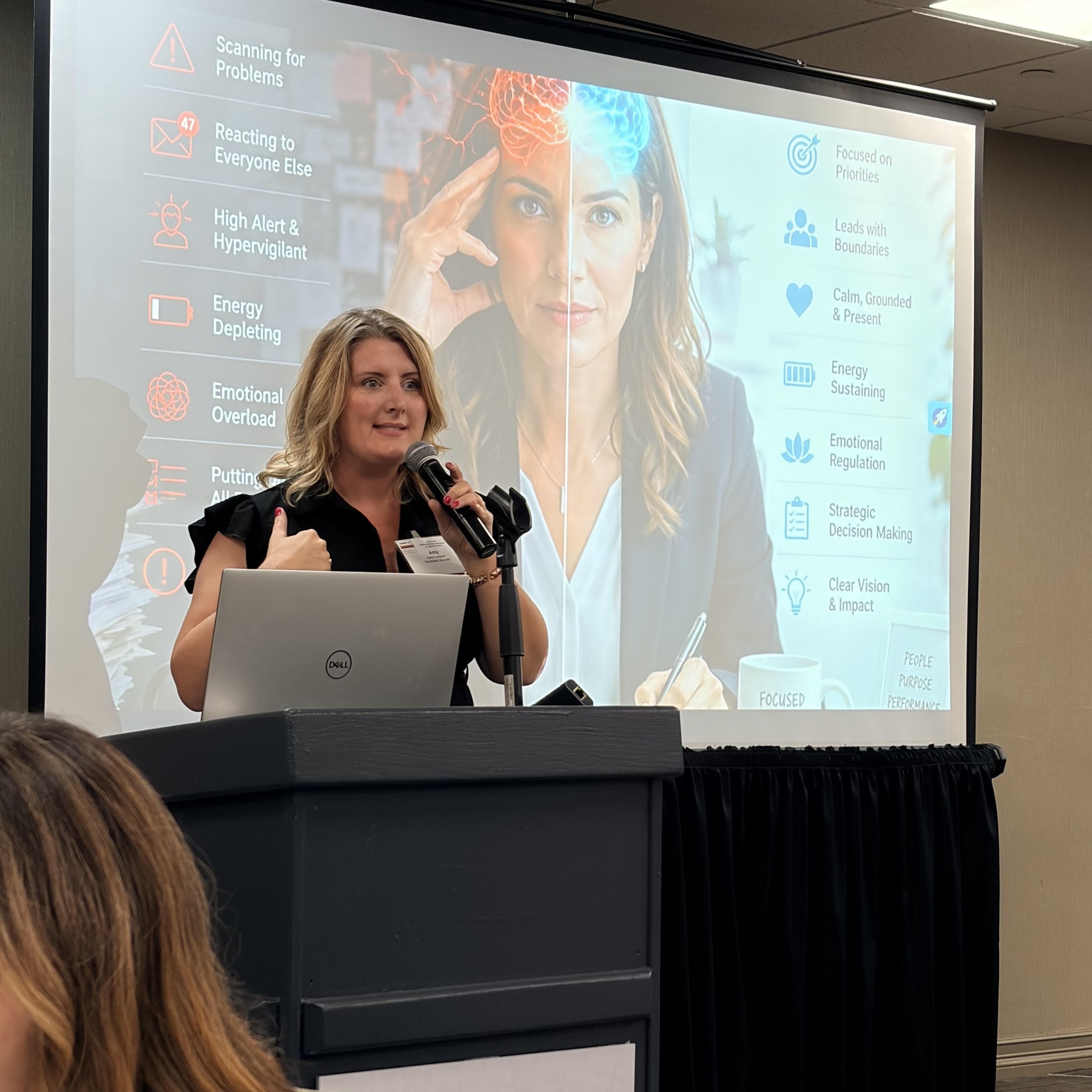 A woman with blonde hair and a black top speaking into a microphone in front of a projection screen displaying a presentation slide.
