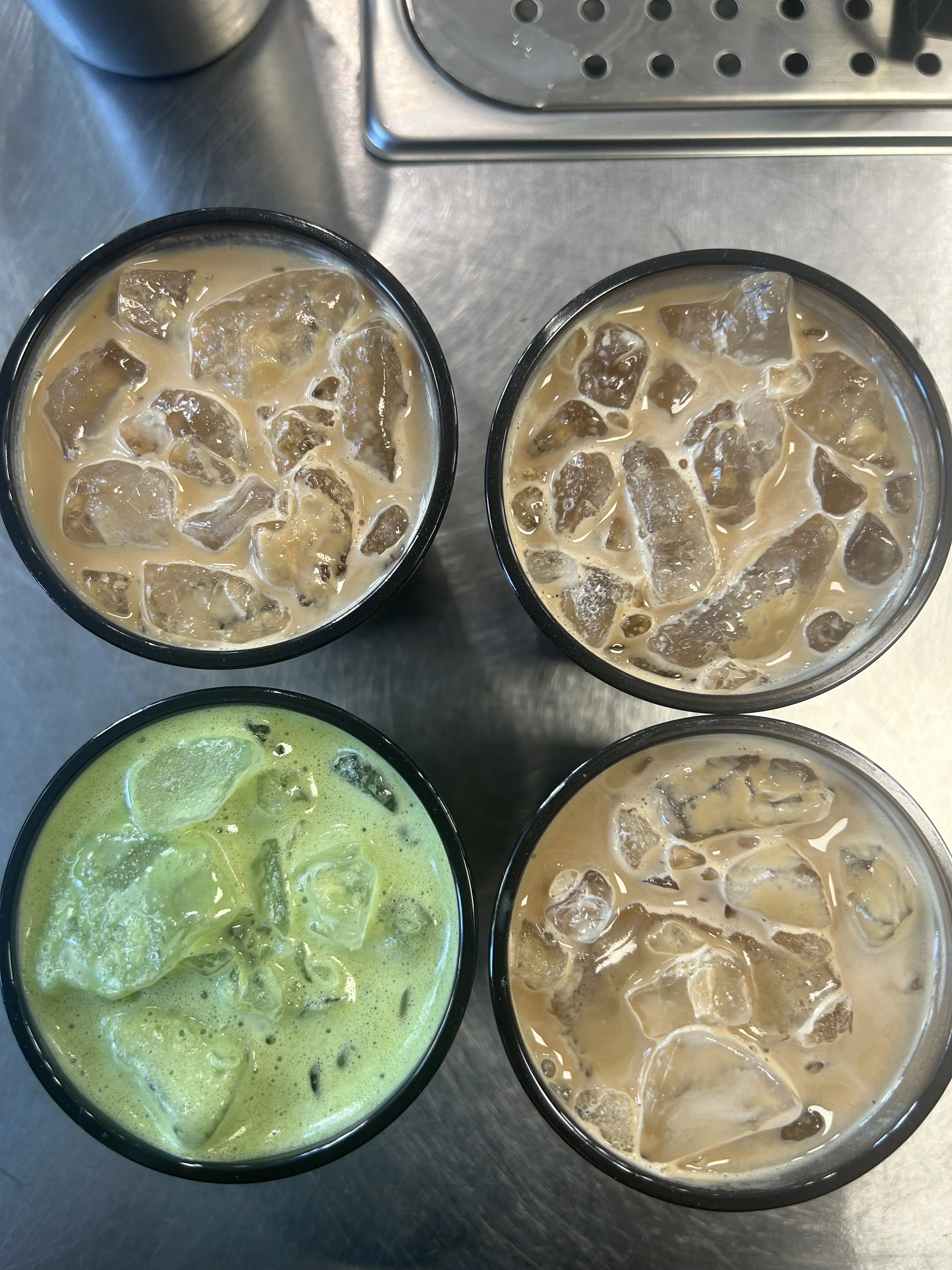 Two glasses of iced coffee and a glass of iced matcha latte on a metal table, with a stainless steel countertop in the background.