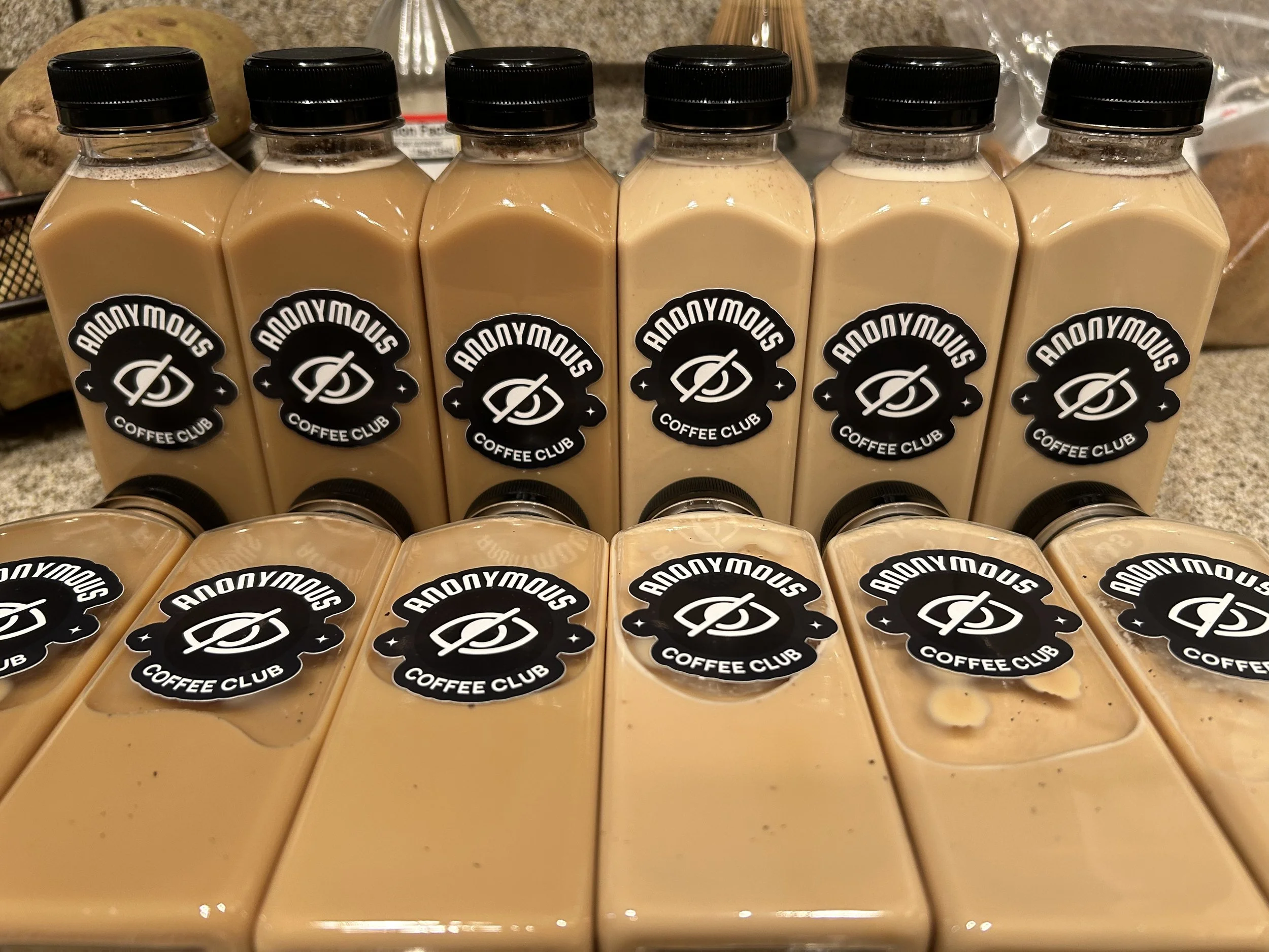 Six bottles of coffee drink lined up on a counter, each with a black cap and a black sticker labeled "Anonymous Coffee Club" with a logo.