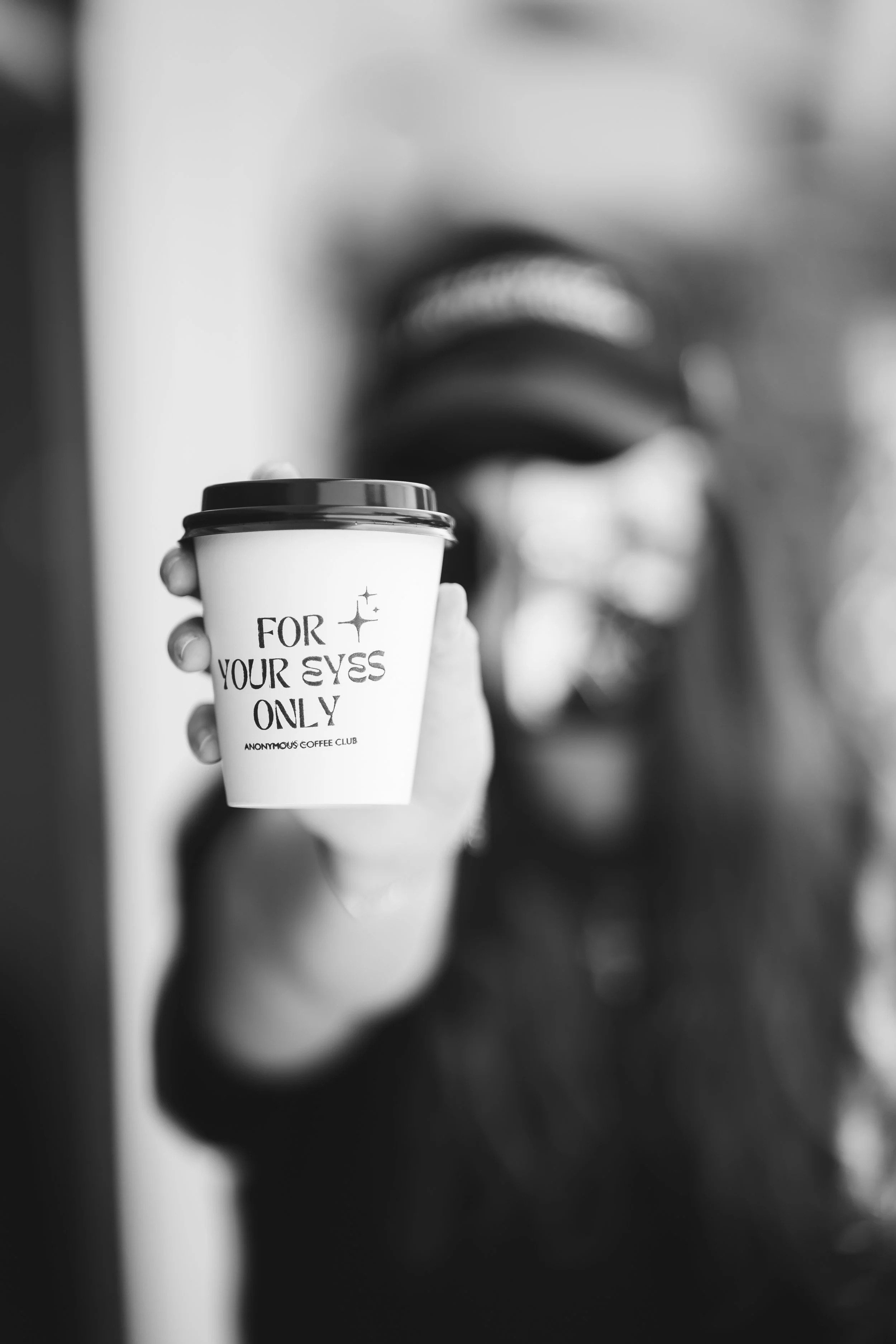 Person holding a coffee cup with the words 'For your eyes only' written on it, in focus. The person is smiling, and the background is blurred.