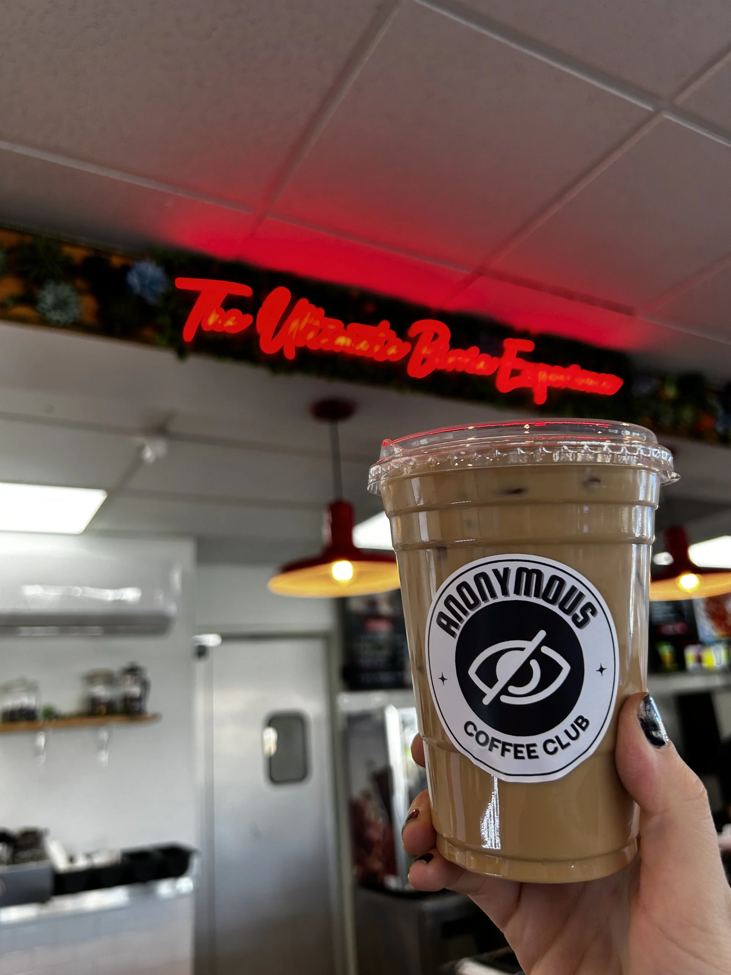 A hand holding an iced coffee cup with a logo that says 'Anonymous Coffee Club' inside a coffee shop with a rear wall that has a neon sign and shelves.