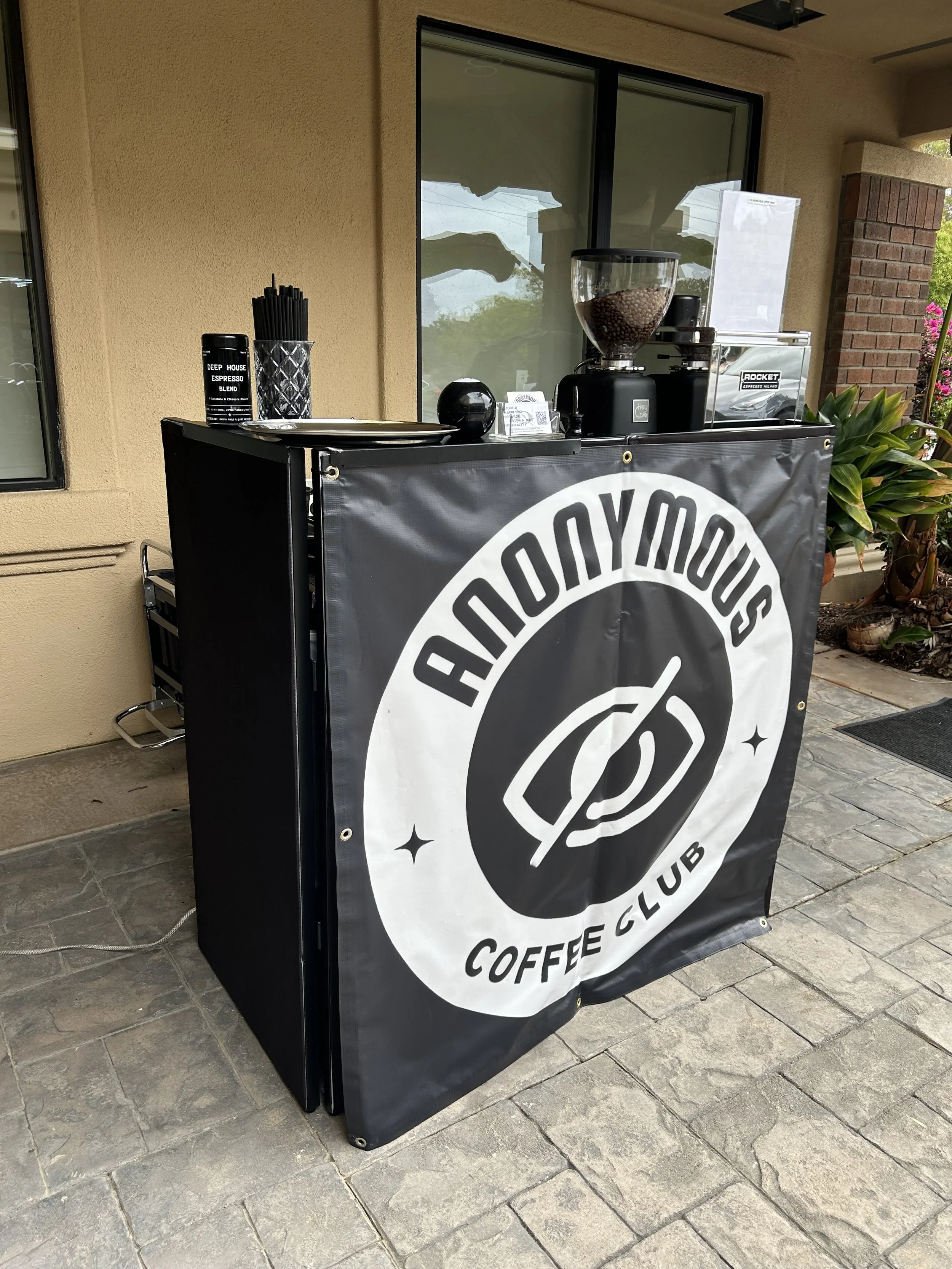 A coffee station on a black cart featuring a coffee banner, a coffee grinder with coffee beans, a container of black straws, and a coffee machine, set outside a building with beige walls and a window.