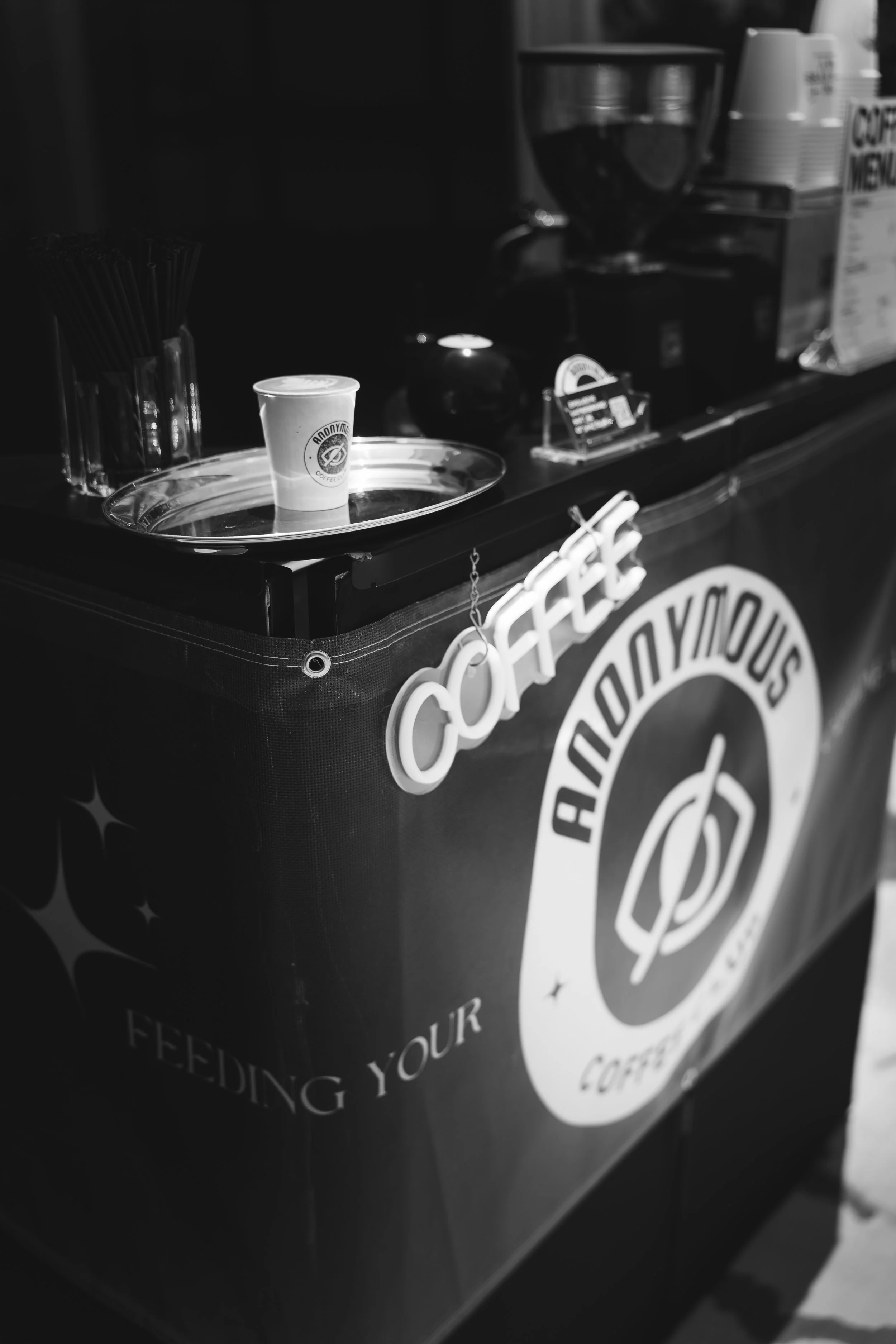 A black and white photo of a coffee cart with a sign reading "Coffee" in large letters. On top of the cart is a tray with a single paper coffee cup featuring a logo, and in the background, there are coffee equipment and stacks of cups.