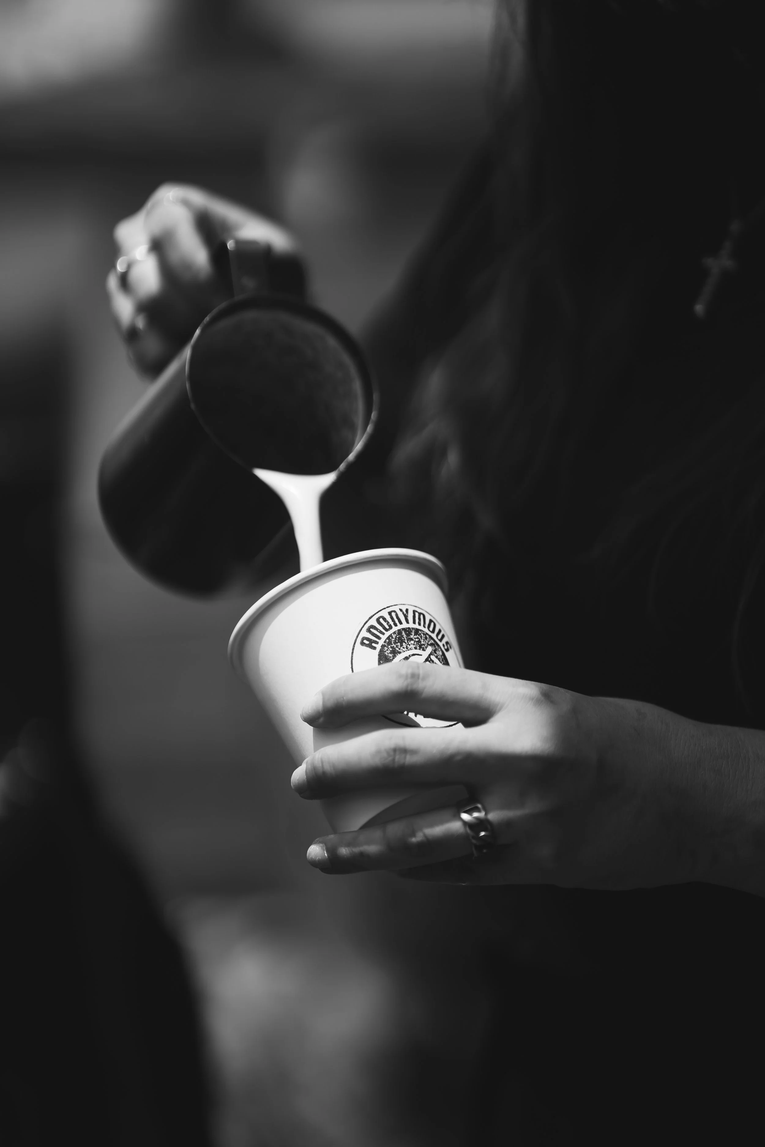 Close-up of a person pouring a drink from a dark bottle into a disposable cup, with the logo "ANONYMOUS" visible on the cup. The person has multiple rings on their fingers and a bracelet with a cross.