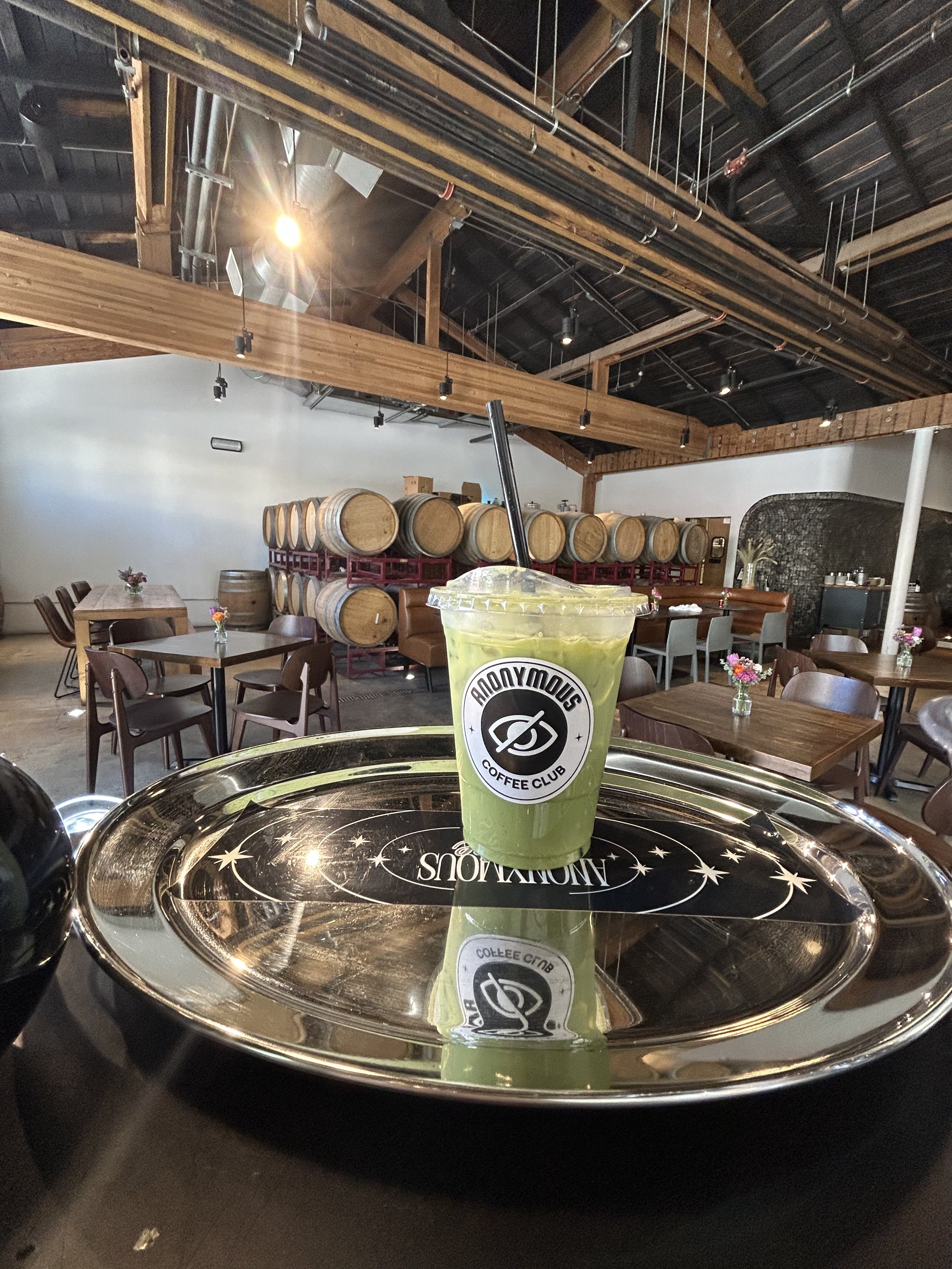 A green beverage in a clear plastic cup with a straw, placed on a stainless steel tray that reflects the cup. The tray has a design with stars and the word 'MOONLIGHT.' The background shows a room with wooden barrels, tables, chairs, and a ceiling with exposed pipes and wood beams, indicating a brewery or taproom setting.