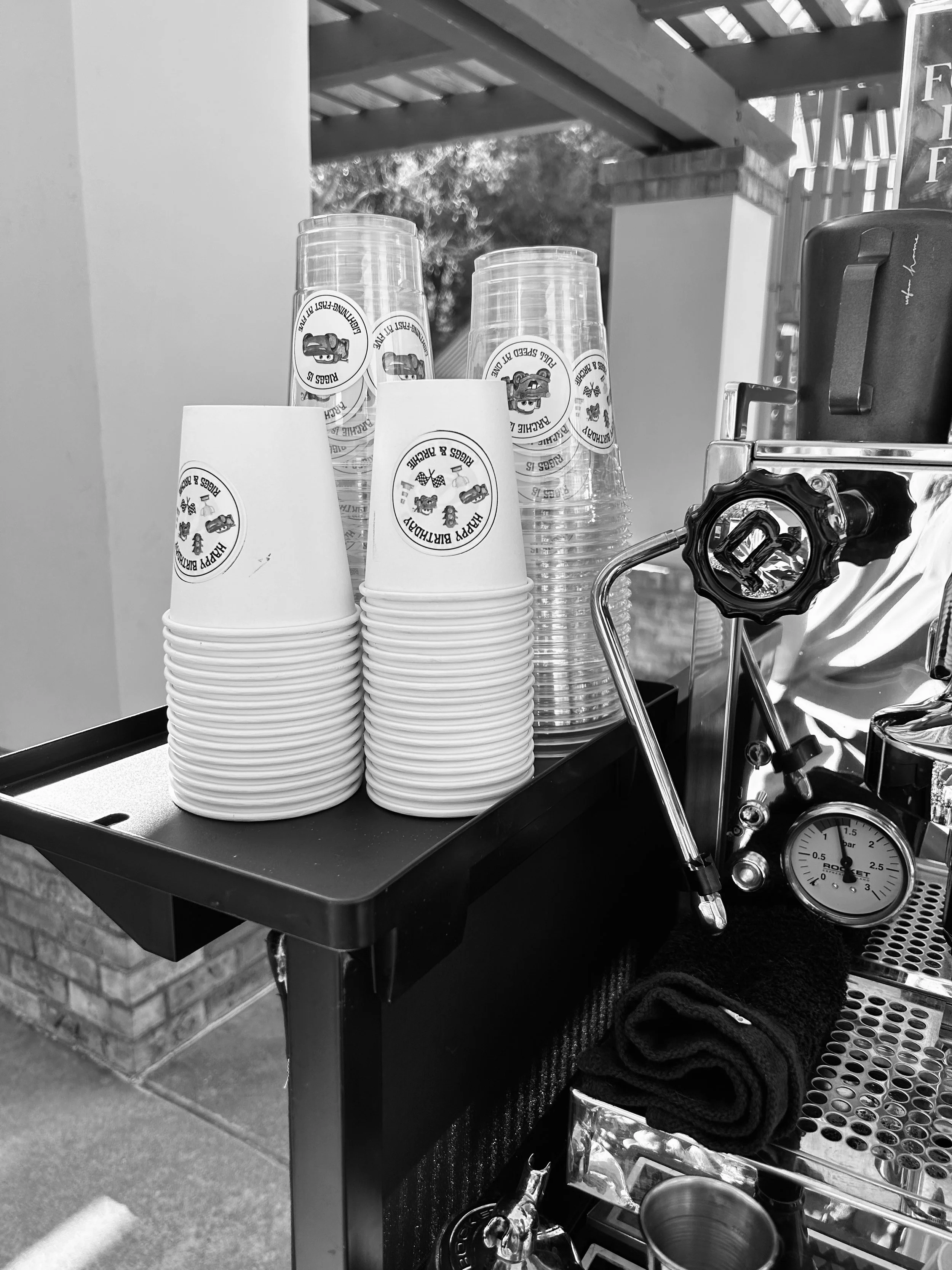 Stacked paper cups with a logo, a coffee machine, and a black towel on a black serving cart outside.