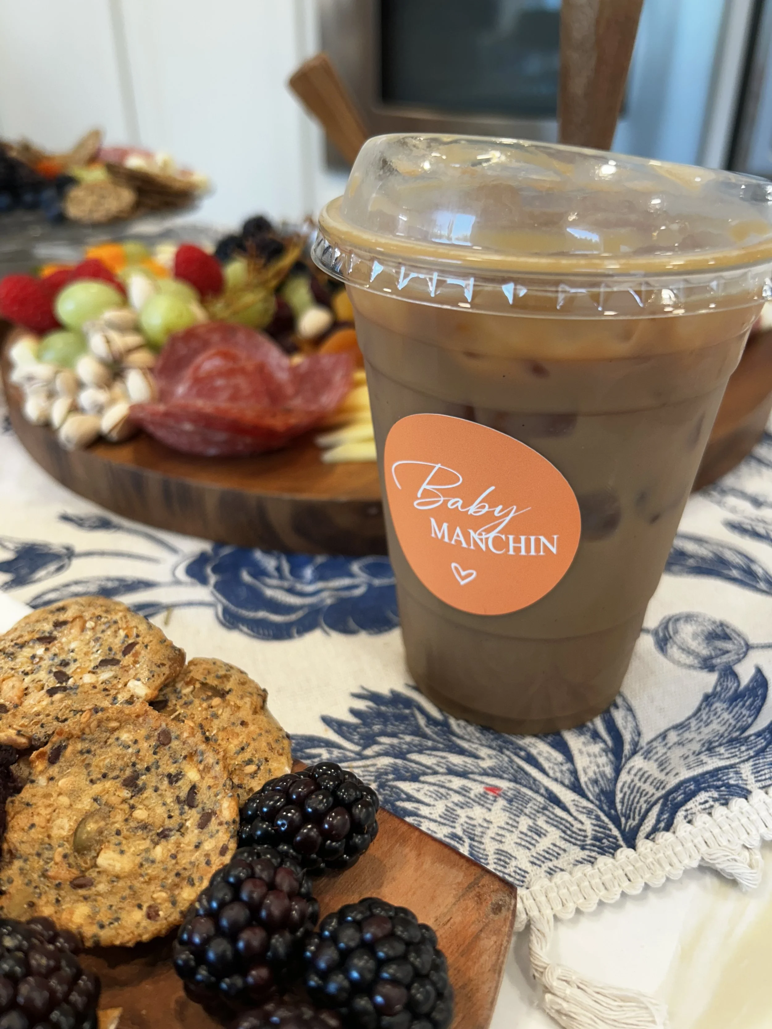 A close-up of a plastic cup of iced coffee with a clear lid, placed on a table with cookies, blackberries, and a tray of colorful fruit in the background.