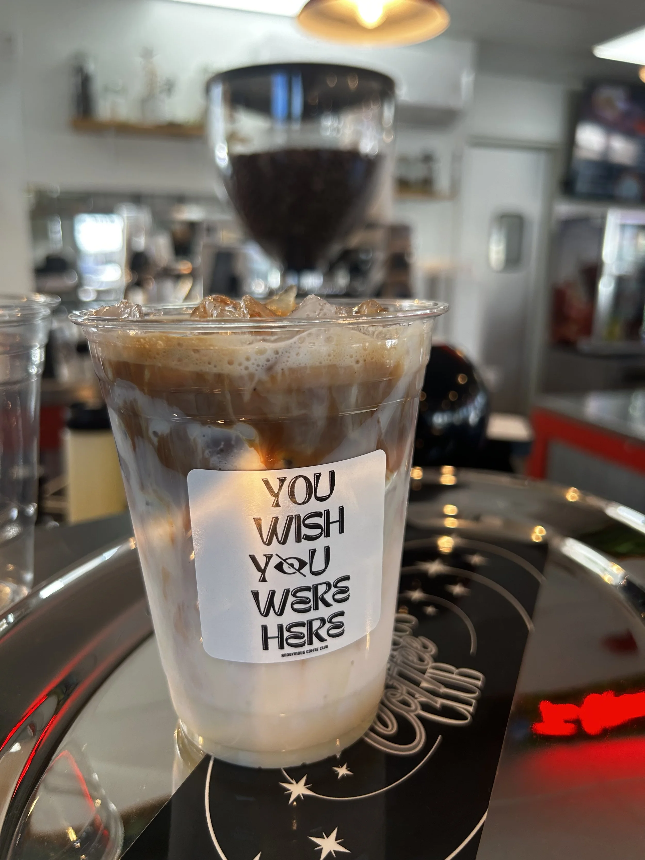 Close-up of a tall glass filled with iced coffee and cream, sitting on a stovetop with a blurred background of a kitchen. The glass has a label with the phrase 'YOU WISH YOU WERS HERS' written in black. Behind the glass, a coffee grinder with dark coffee beans is visible.