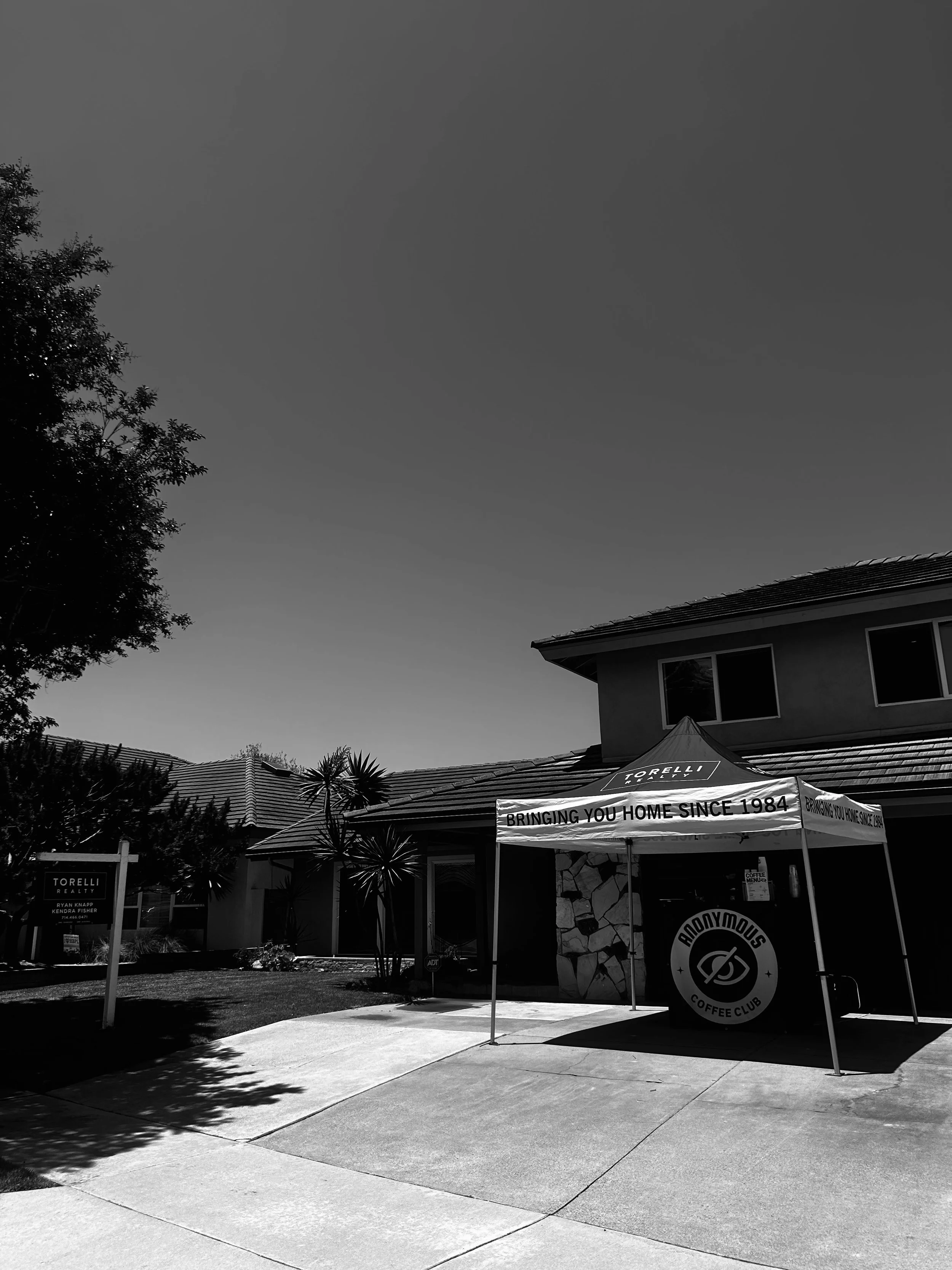A building with a banner stating 'BRINGING YOU HOME SINCE 1984' in front of a coffee club tent. There are trees, bushes, and a sidewalk in front of the building.