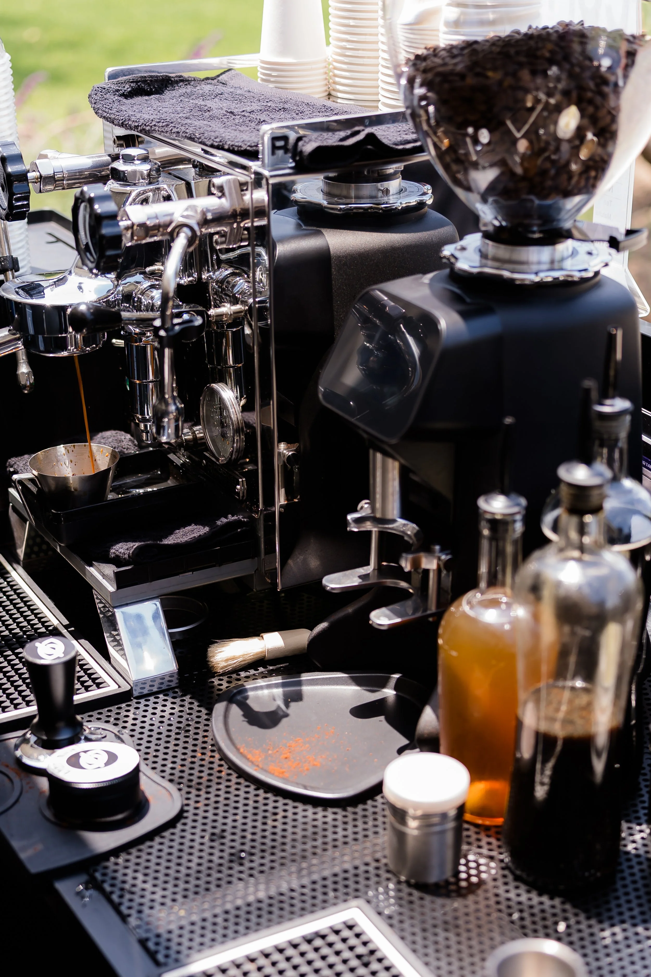 A coffee station with an espresso machine, coffee beans, cups, and bottles of flavored syrup on a black counter with a metal surface.