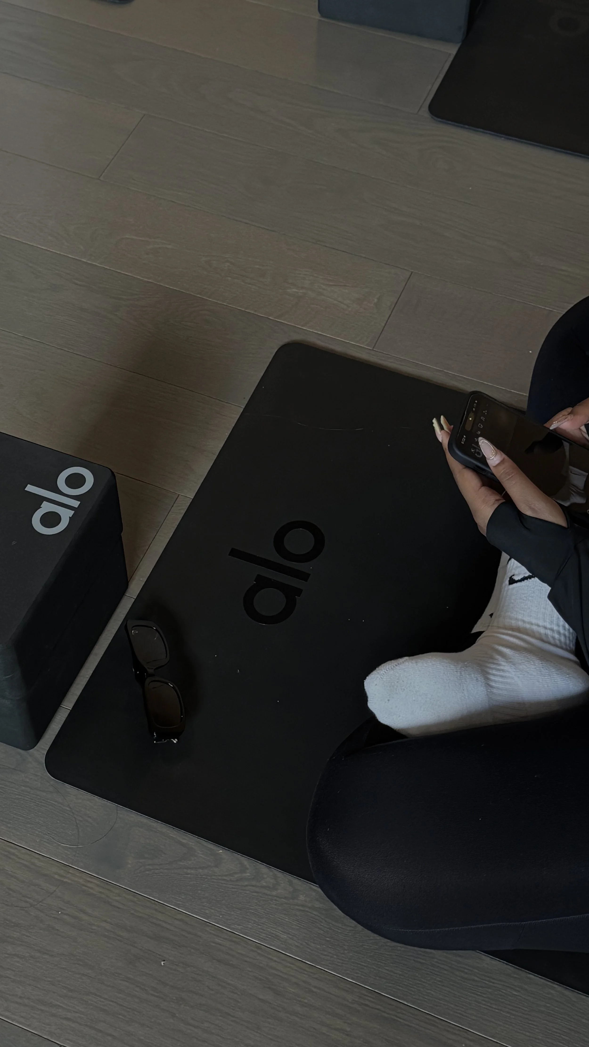 Person sitting on a black yoga mat with the word 'alo' on it, using a smartphone, with sunglasses on the mat, and a yoga block with 'alo' on it nearby, against a wooden floor background.