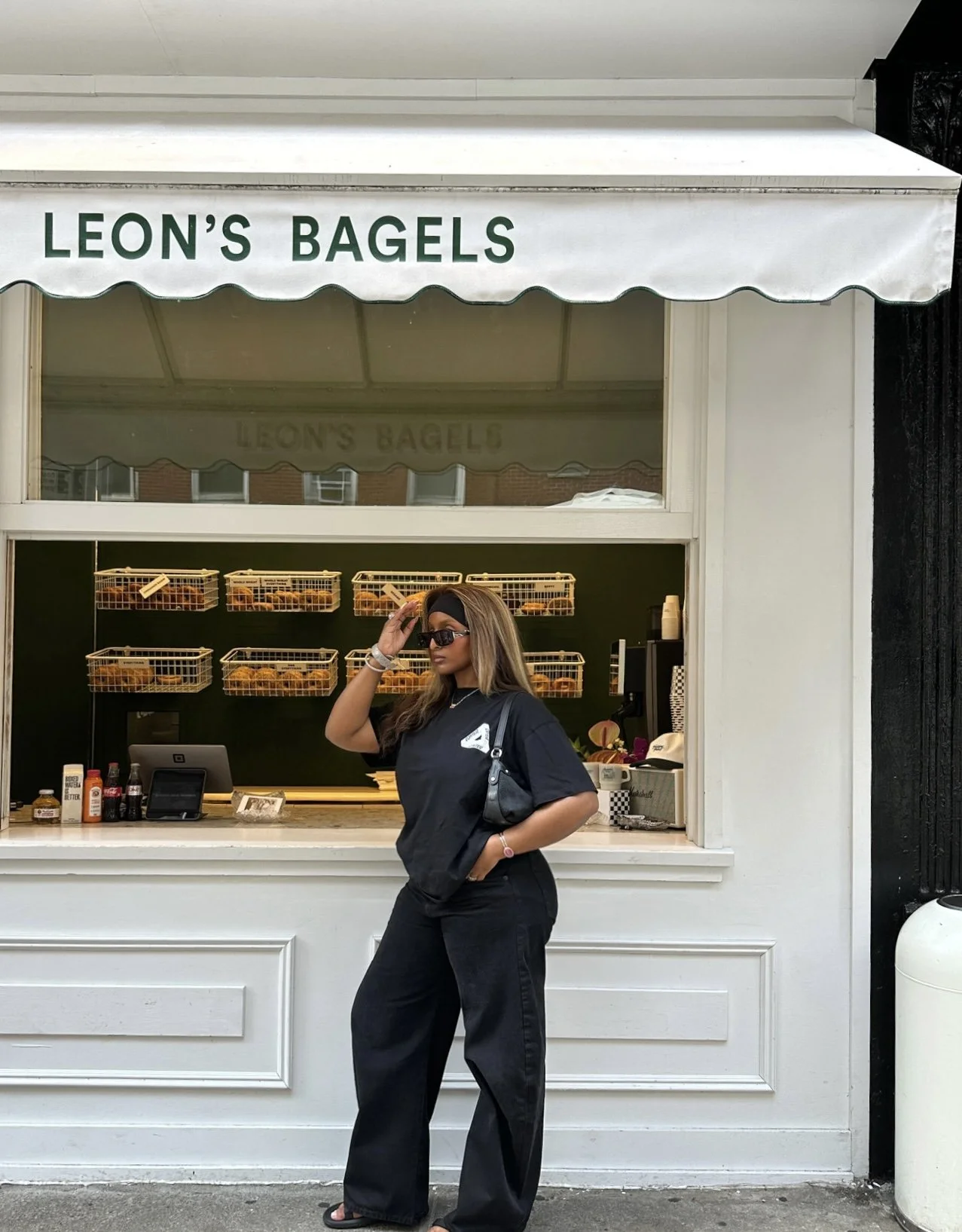A woman wearing black Dezi sunglasses and black Corteiz t-shirt standing outside a bagel shop called 'Leon's Bagels'. The shop has a white awning with green text and a window display with baskets of bagels.