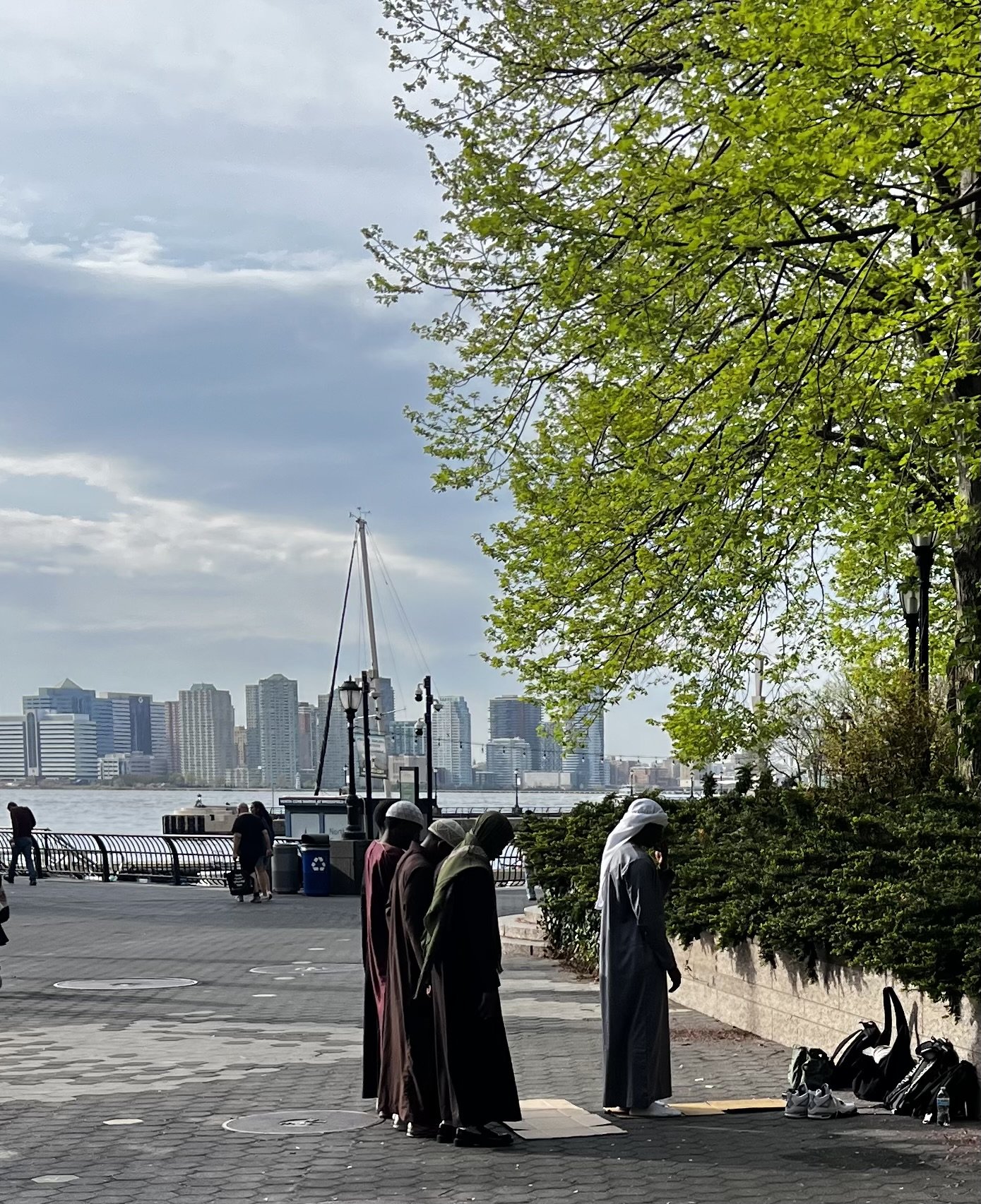 Five Muslim men praying along a riverside promenade with New York's skyline in the background, some wearing traditional robes and head coverings, shoes and backpacks beside them, under green leafy trees.
