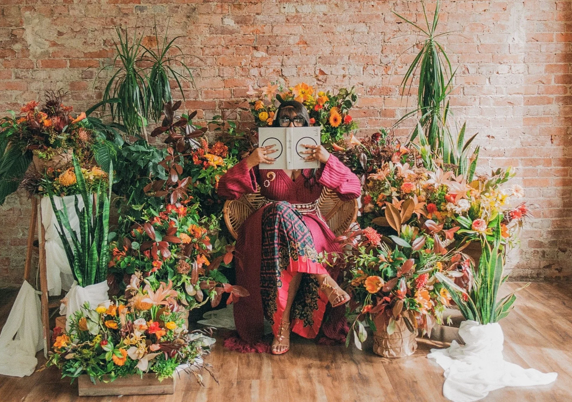 A woman wearing a red and pink patterned dress and glasses sitting on a rattan chair, surrounded by a large arrangement of colorful flowers and green plants against a brick wall.