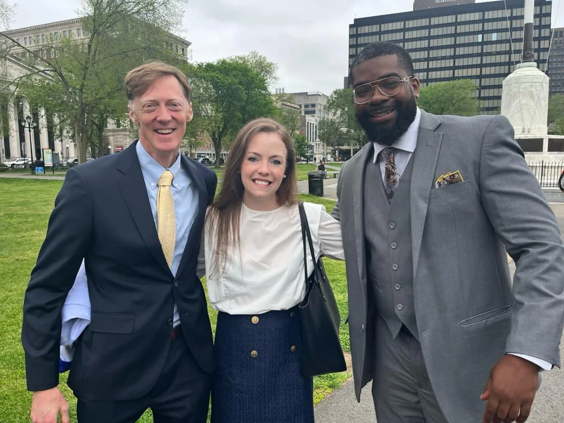 Three people smiling outdoors in a park, dressed in business attire, with city buildings and trees in the background.
