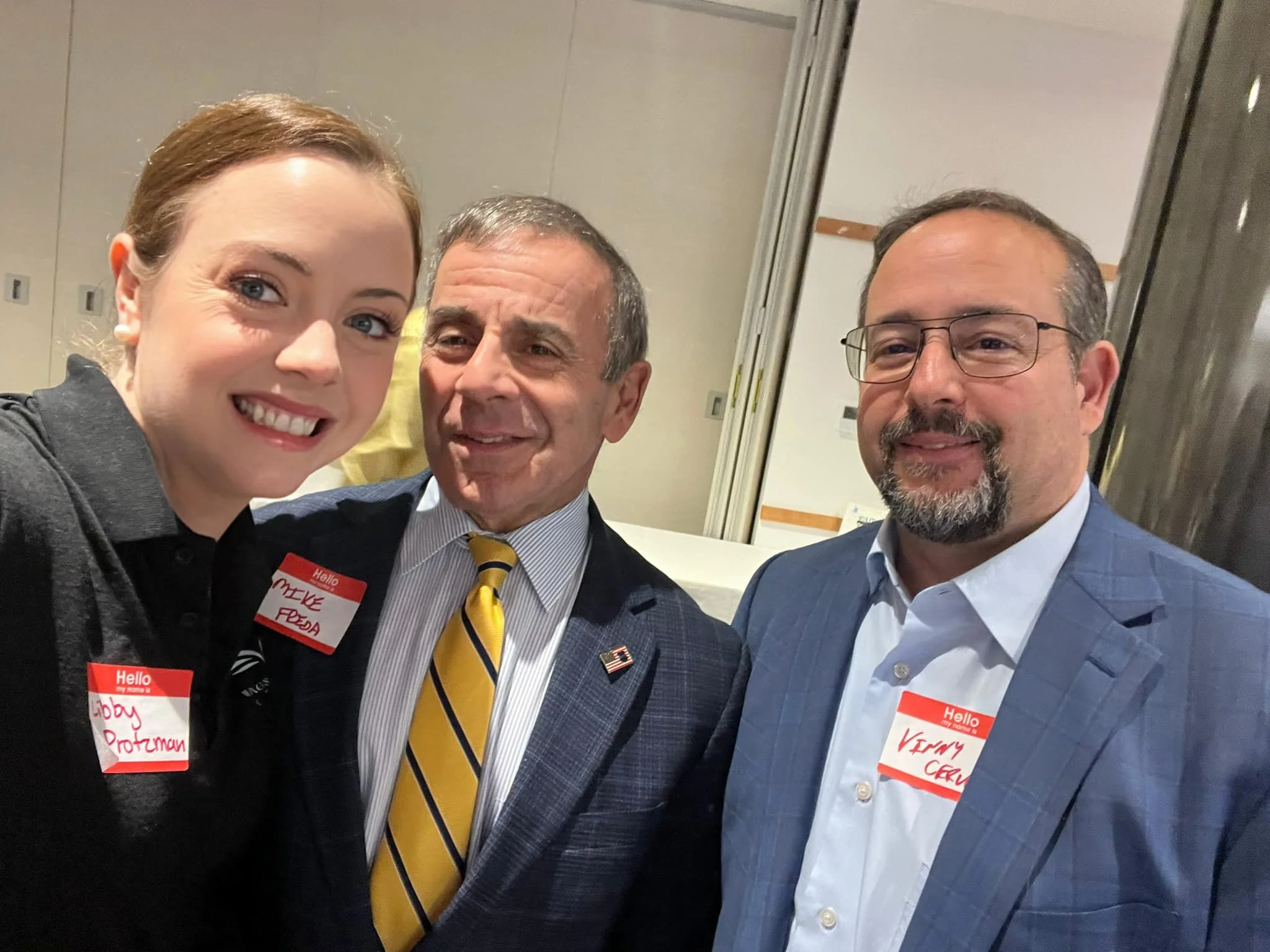 Three people standing close together at an indoor event, smiling for a photo. They are wearing name tags; the woman on the left has a bald head and is wearing a black shirt, the man in the middle has gray hair and a striped yellow tie, and the man on the right has glasses and a gray suit with a light blue shirt.