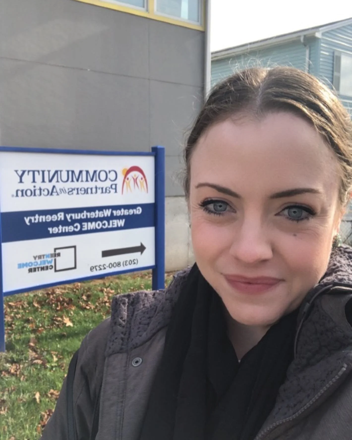Selfie of a woman with blue eyes and brown hair taking a picture outside near a sign for the community partnership and weather refuge center.