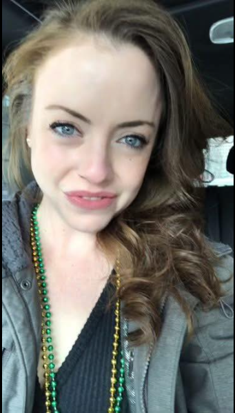 A young woman with long, wavy, reddish-brown hair and blue eyes taking a selfie. She is smiling slightly and wearing a gray jacket over a dark top, with green and gold beads around her neck. The background shows parts of a car's interior.