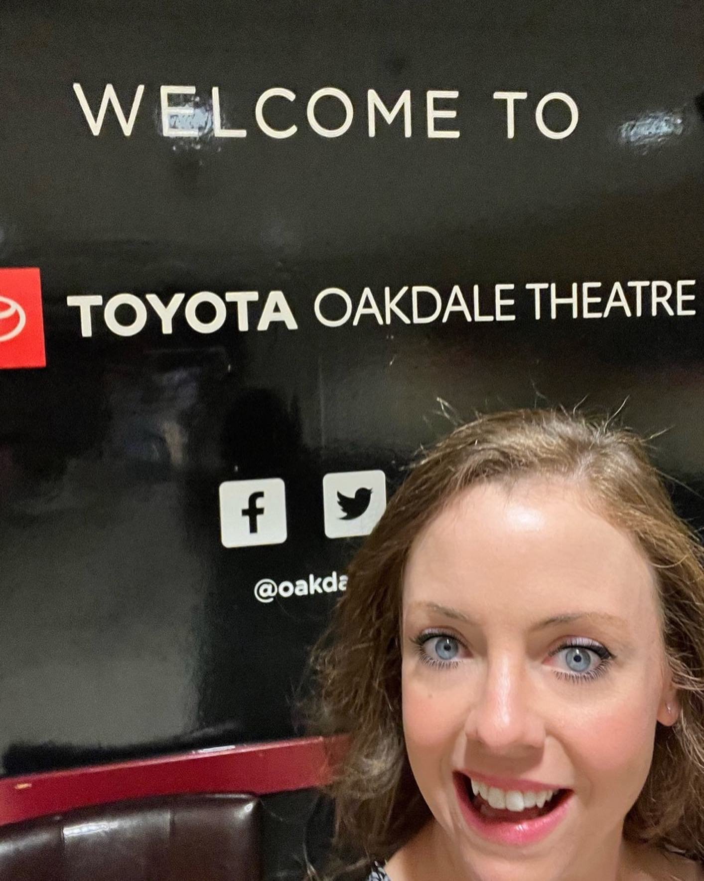 Woman with curly hair and blue eyes smiling in front of a sign that reads 'Welcome to Toyota Oakdale Theatre,' with social media icons for Facebook and Twitter.