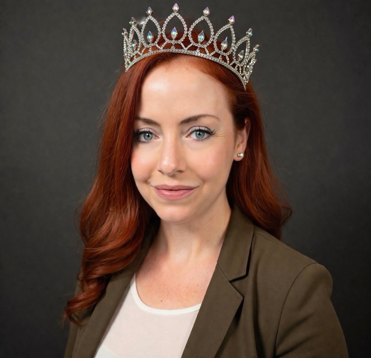 A woman with red hair wearing a tiara and a brown blazer on a dark background.