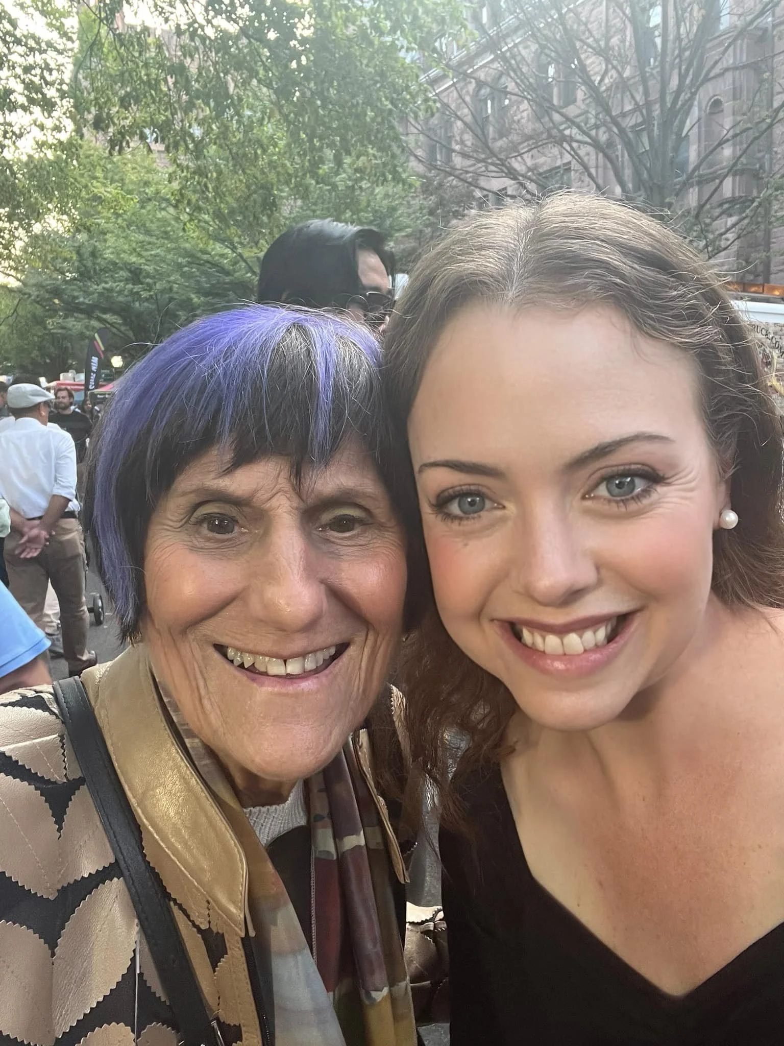 A woman and a younger woman taking a selfie outdoors at a crowded event with trees and buildings in the background.