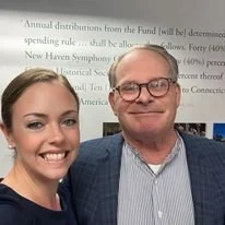 A young woman and an older man smiling for a selfie in front of a projected presentation or document