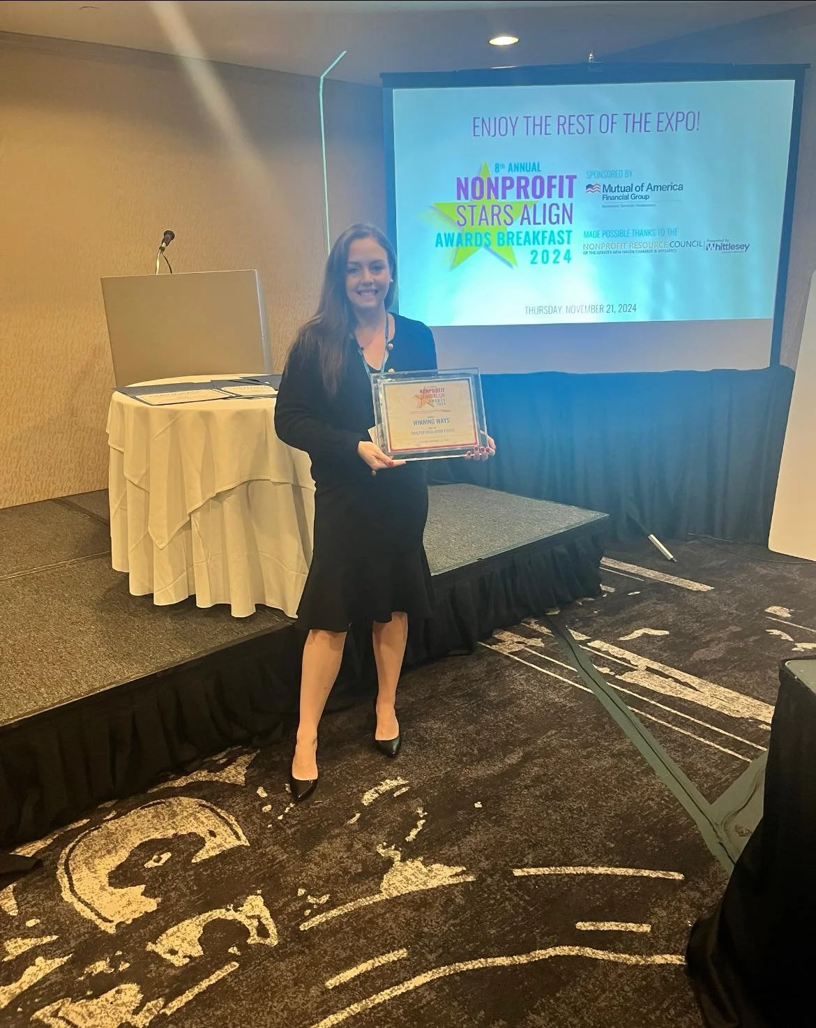 A woman in a black dress and blazer holding an award certificate on stage. Behind her is a presentation screen with event details for the 8th Annual Nonprofit Stars Align Awards Breakfast 2024, sponsored by Mutual of America Financial Group, held on November 21, 2024.