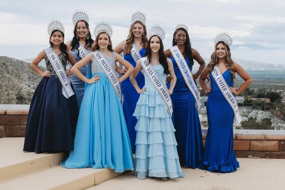 Seven young women wearing crowns and sashes standing outdoors, dressed in blue gowns, with a scenic landscape in the background.