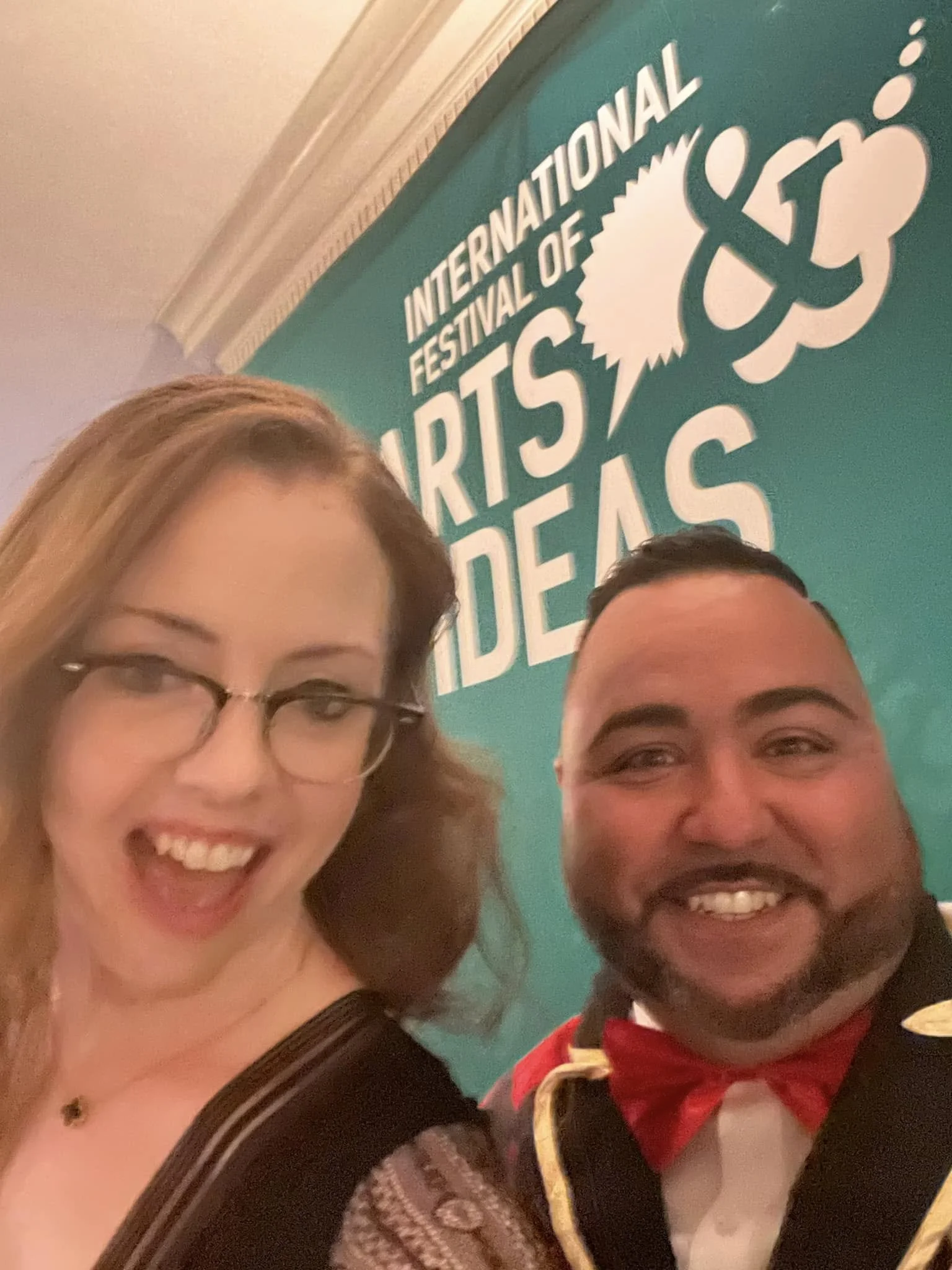 Two smiling people taking a selfie at the International Festival of Arts & Ideas, with a teal-colored sign in the background.