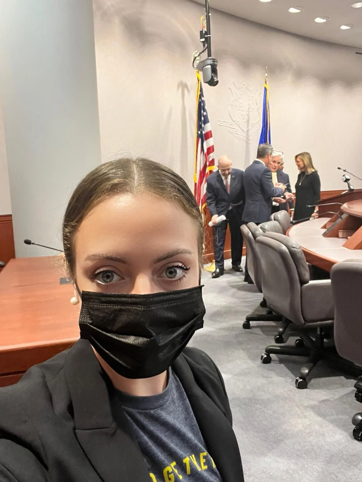 A woman wearing a black face mask, standing in a conference room with several people in formal attire, American flags, and a seal on the wall behind them.