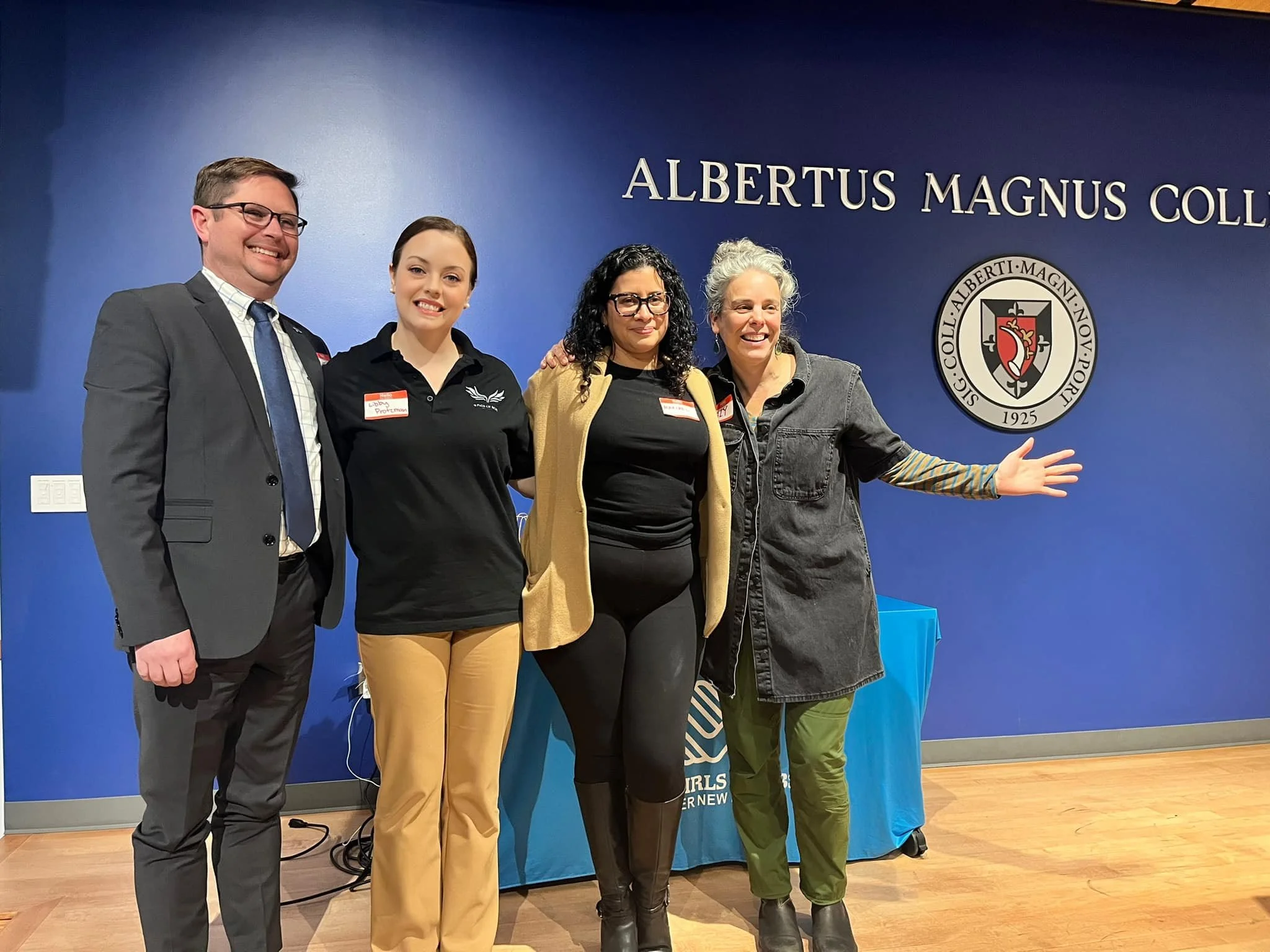 Group of four people standing together in front of a blue wall with the words 'ALBERTUS MAGNUS COL...' and a college seal, posing for a photo with one person gesturing with open arms.