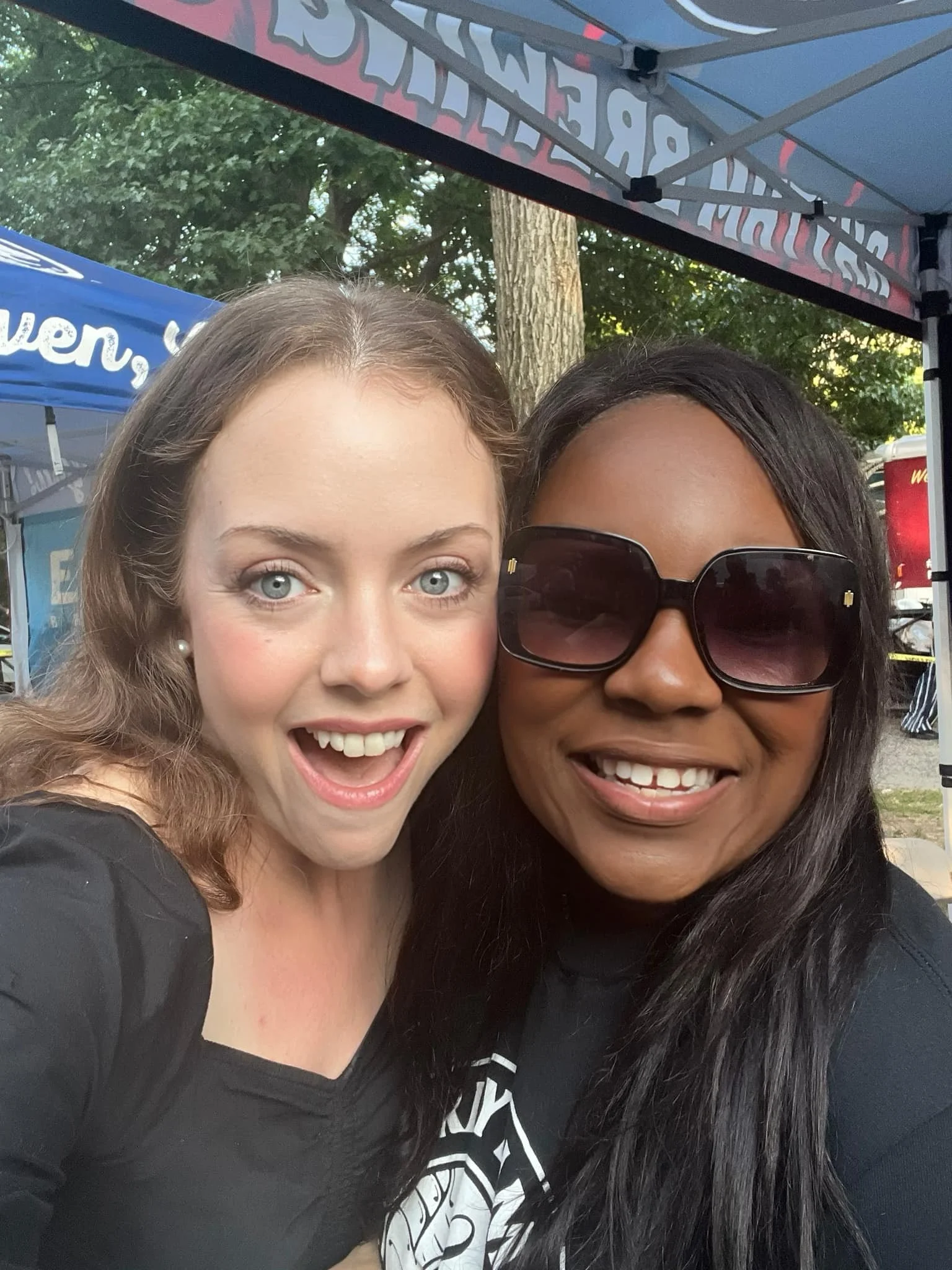 Two women taking a selfie outdoors at a fair or festival. One woman has light skin, blue eyes, and light brown hair, and the other has darker skin, long black hair, and is wearing large sunglasses. They are smiling and appear happy.