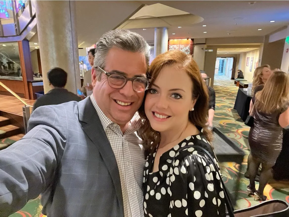 A smiling man and woman taking a selfie at a formal event in a hotel or conference center lobby, with other guests in the background.