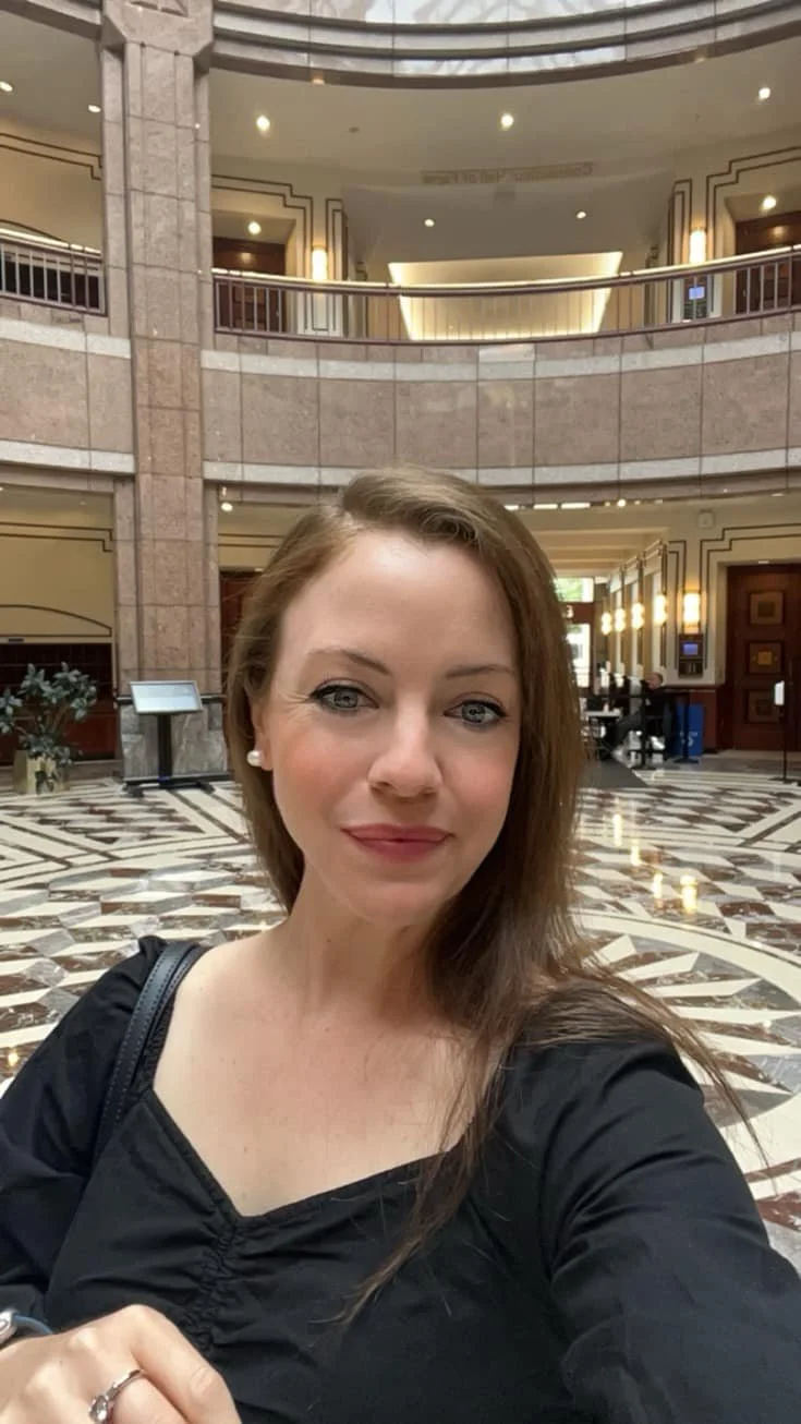 A woman with long brown hair, wearing a black top and pearl earrings, taking a selfie inside a spacious hotel lobby with marble floors, balcony railings, and elegant lighting.