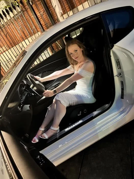 A woman in a white dress sitting in a silver sports car, holding the steering wheel, with a brick wall background.