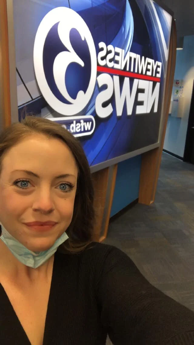 A woman taking a selfie in front of a WTXF FOX television station sign, with her face partially visible and a face mask hanging below her chin.