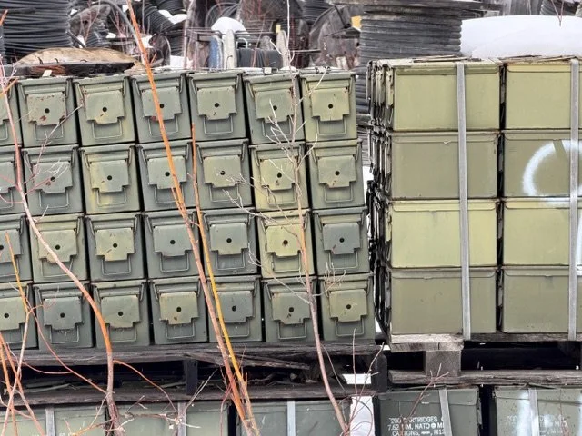 Stacks of green metal ammunition boxes stacked on pallets in an outdoor storage area.