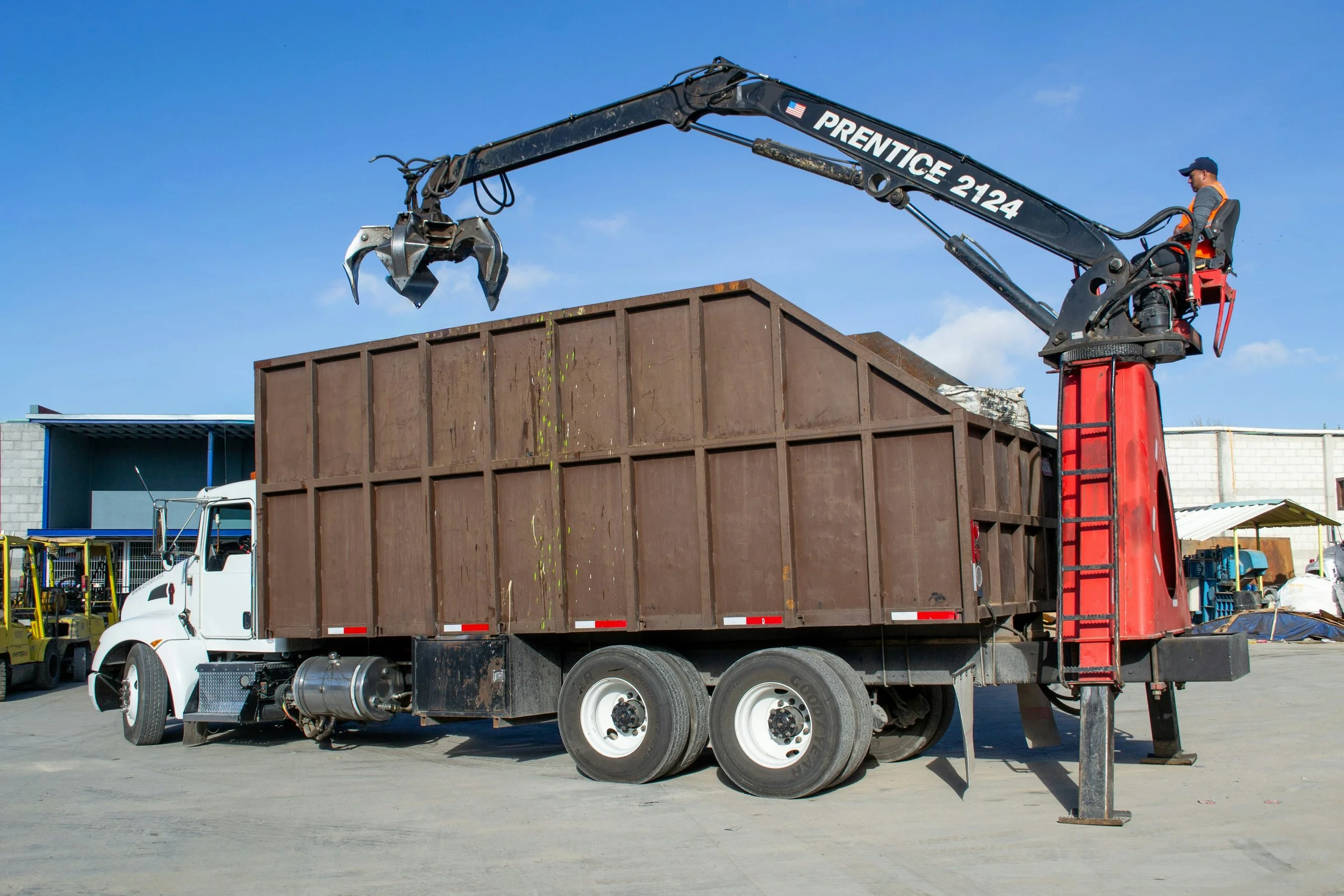 A worker is operating a crane on a flatbed truck, lifting debris into a dumpster on a construction site under a clear blue sky.