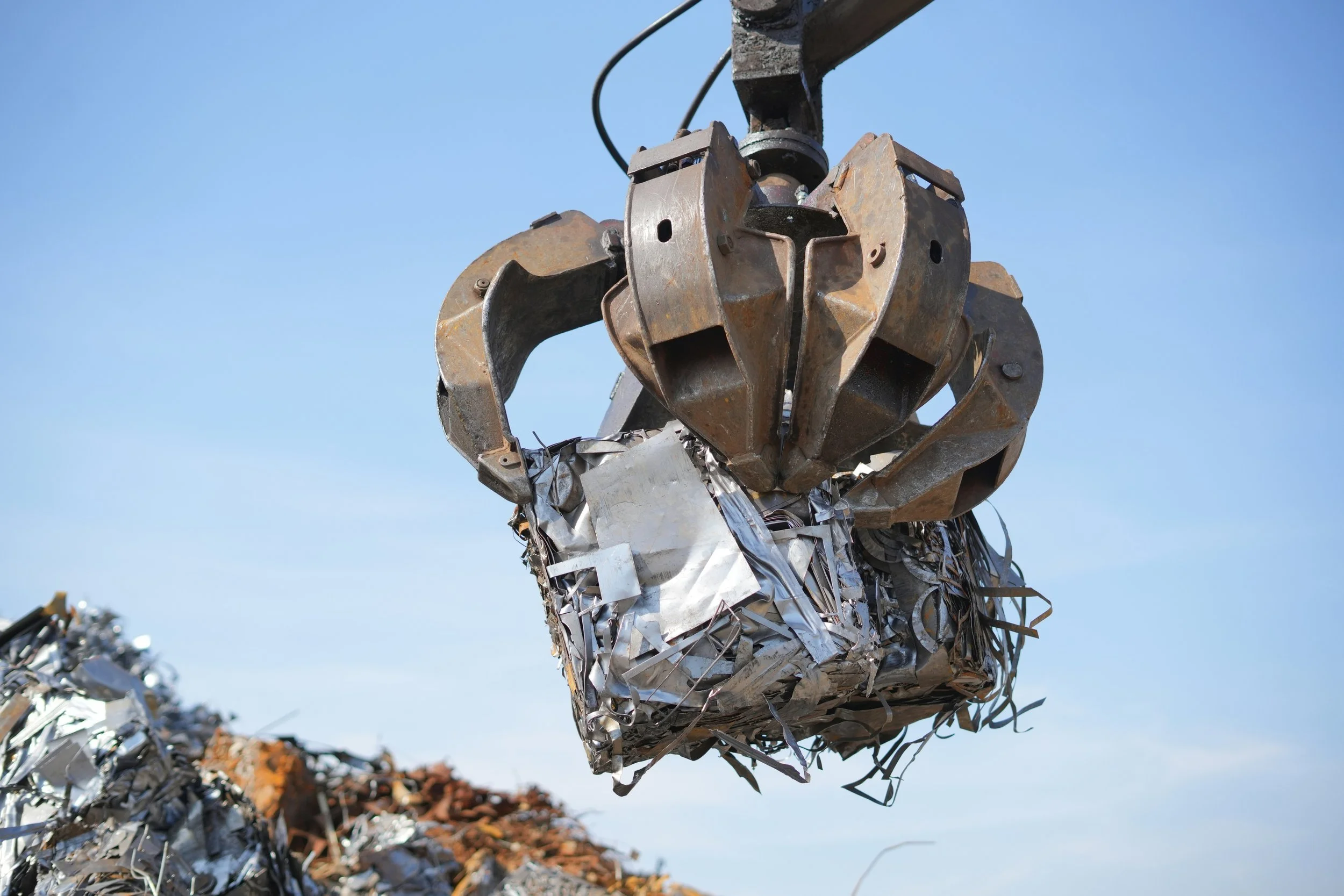 Close-up of a large metal claw lifting crushed aluminum cans against a clear blue sky.