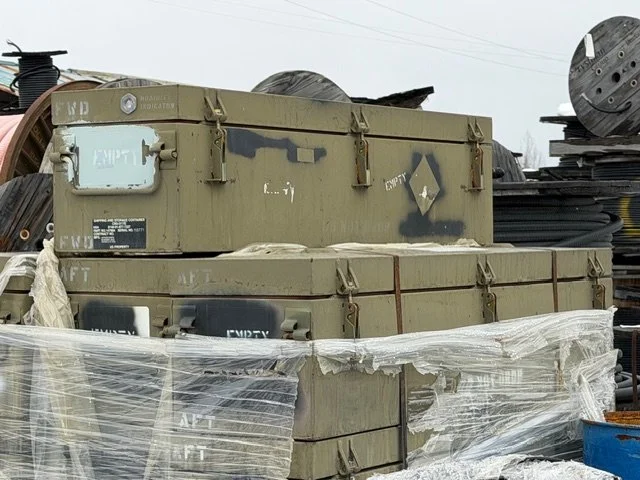 Multiple military-style green metal storage containers stacked outdoors, wrapped in plastic, with spools of cable or wire and other industrial equipment nearby.
