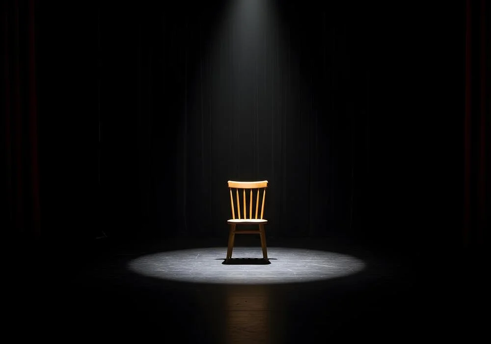 A single wooden chair on a spotlighted stage with a dark background.