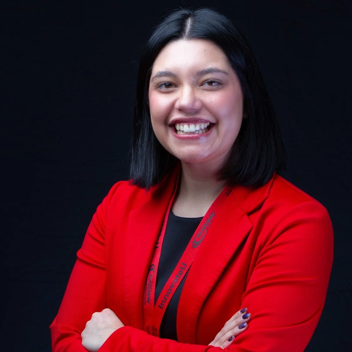 Portrait of a woman with black hair wearing a red blazer and a black top, smiling with crossed arms, against a black background.