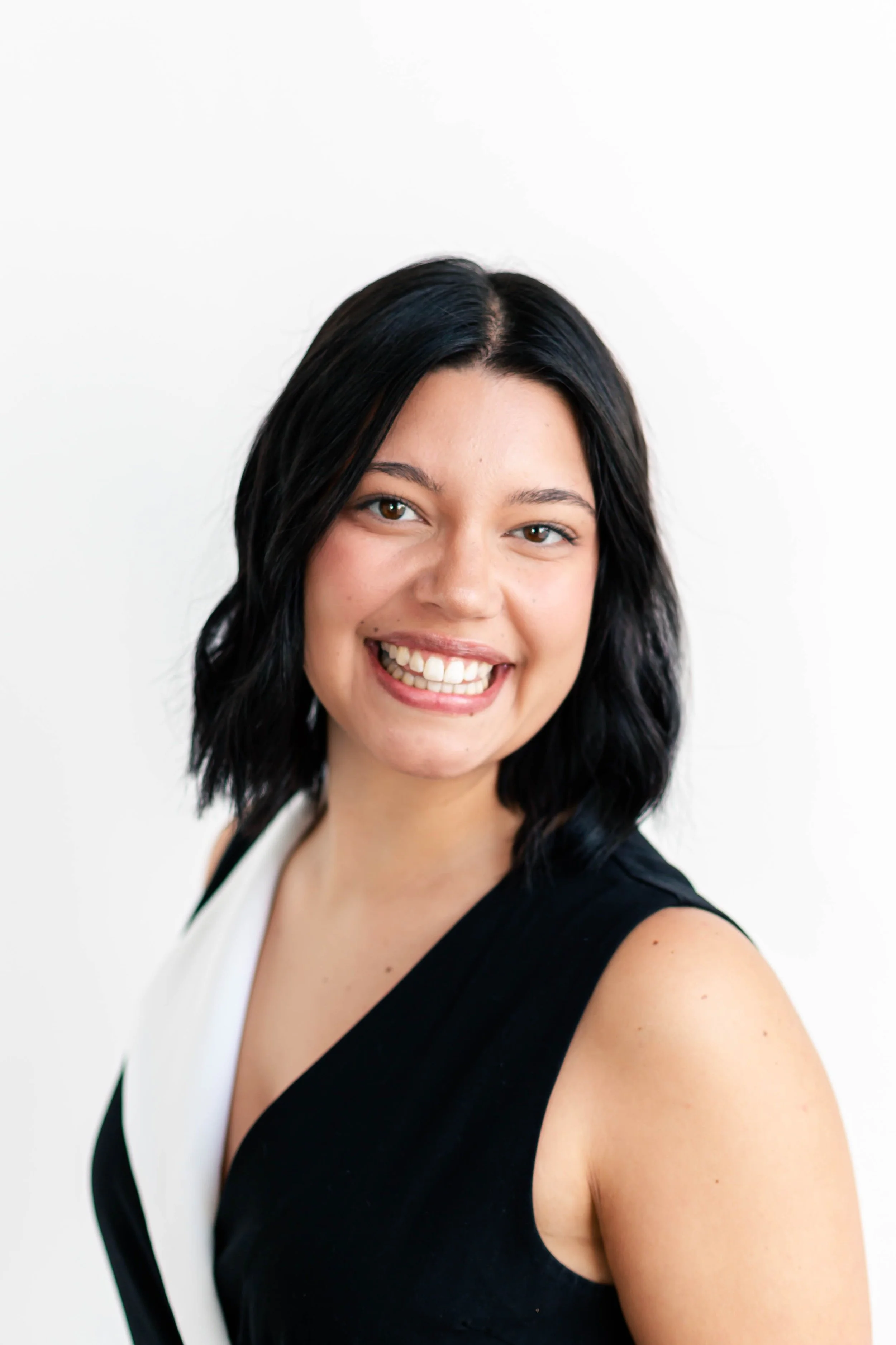 Portrait of a young woman with black, shoulder-length wavy hair, smiling, wearing a sleeveless black and white top, against a plain white background.