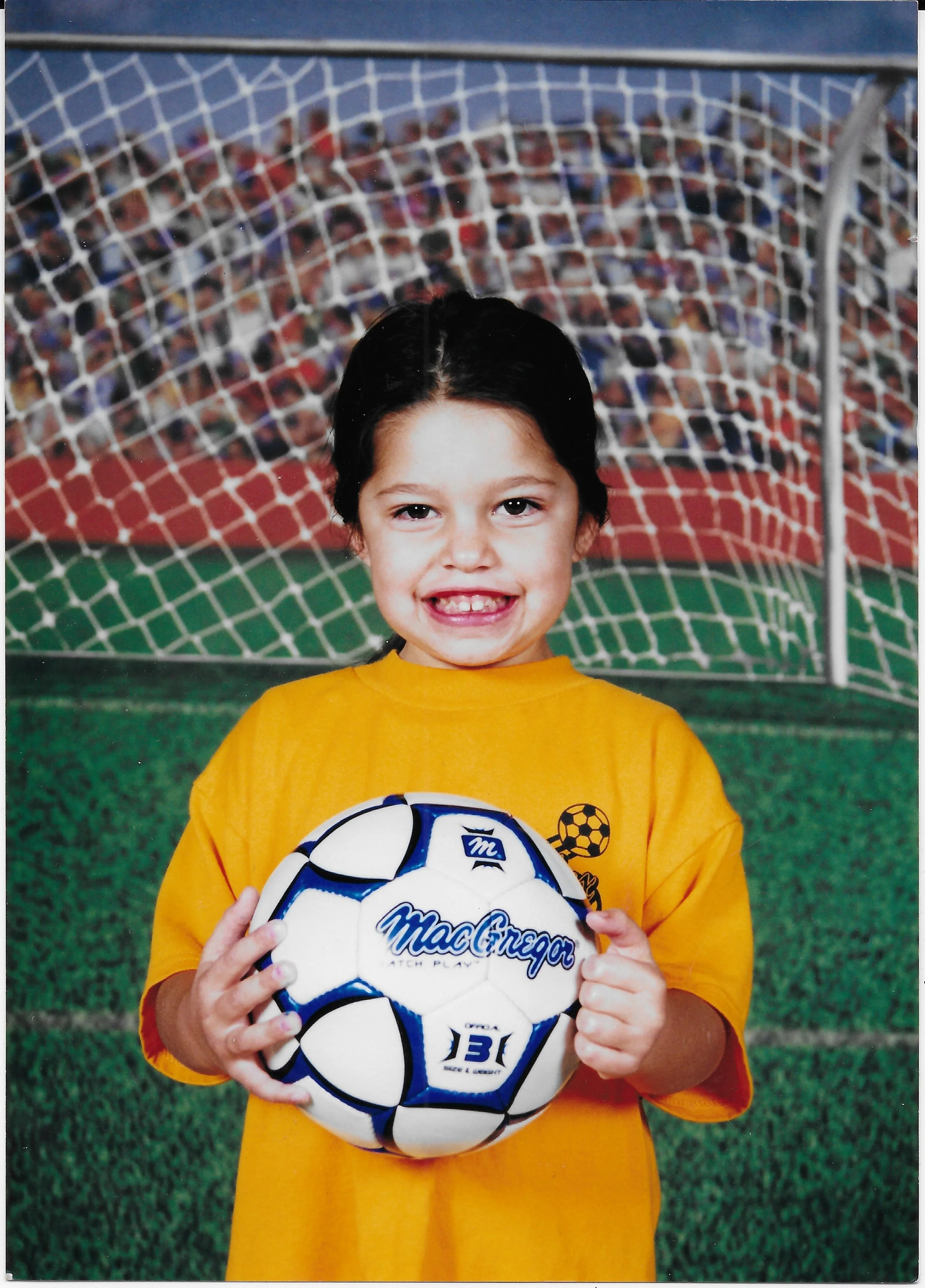 A young girl with dark hair, smiling, holding a soccer ball in front of a goal on a soccer field. She is wearing a yellow jersey and standing on green grass, with a crowded stadium in the background.