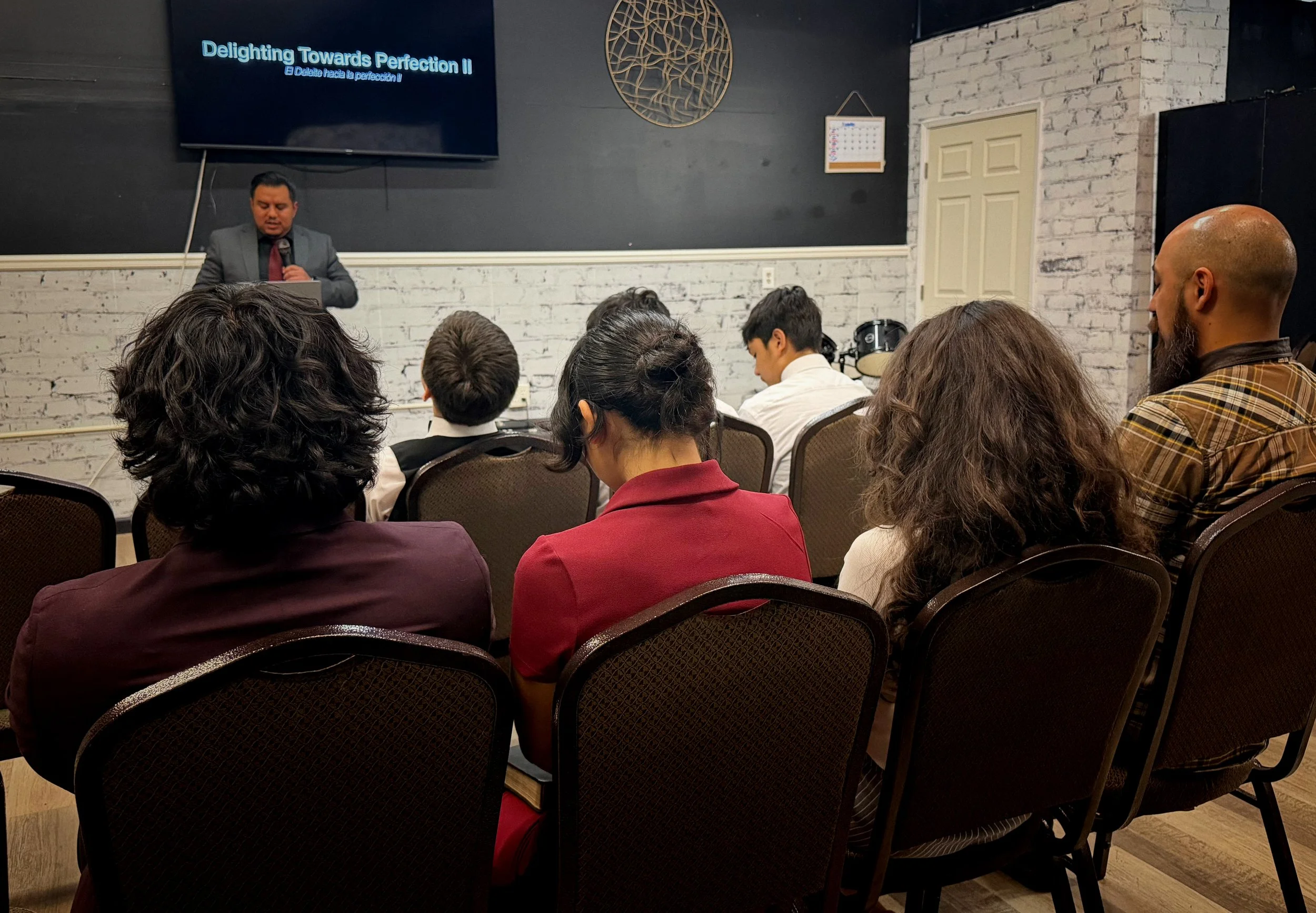 A man in a gray suit and red tie is giving a presentation in front of an audience seated in chairs. The presentation slide on the screen behind him reads 'Delighting Towards Perfection II'. The room has white brick walls and decorative wall art.