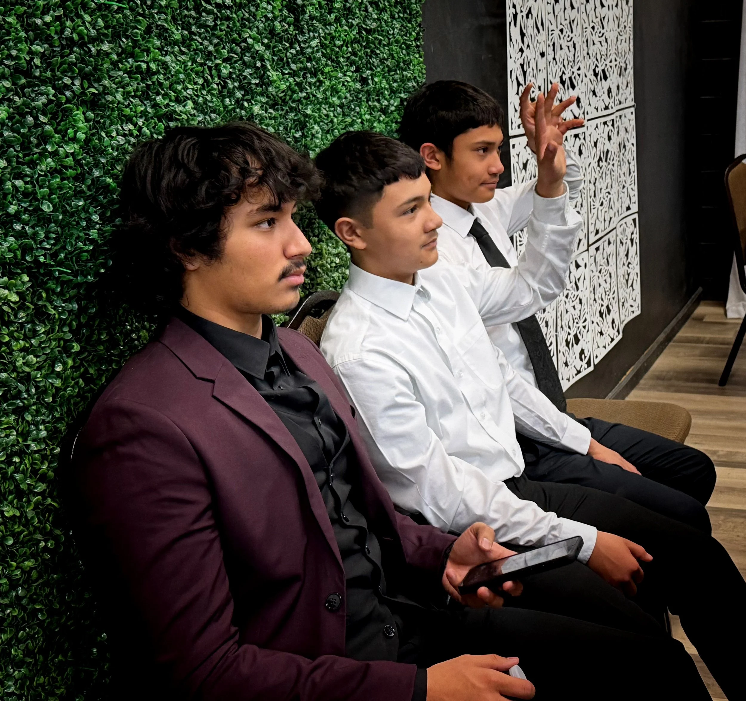 Four young men wearing white shirts and black ties sitting in a row, one is raising his hand. They are seated against a green leafy wall with black and white decorative panels on the right side.